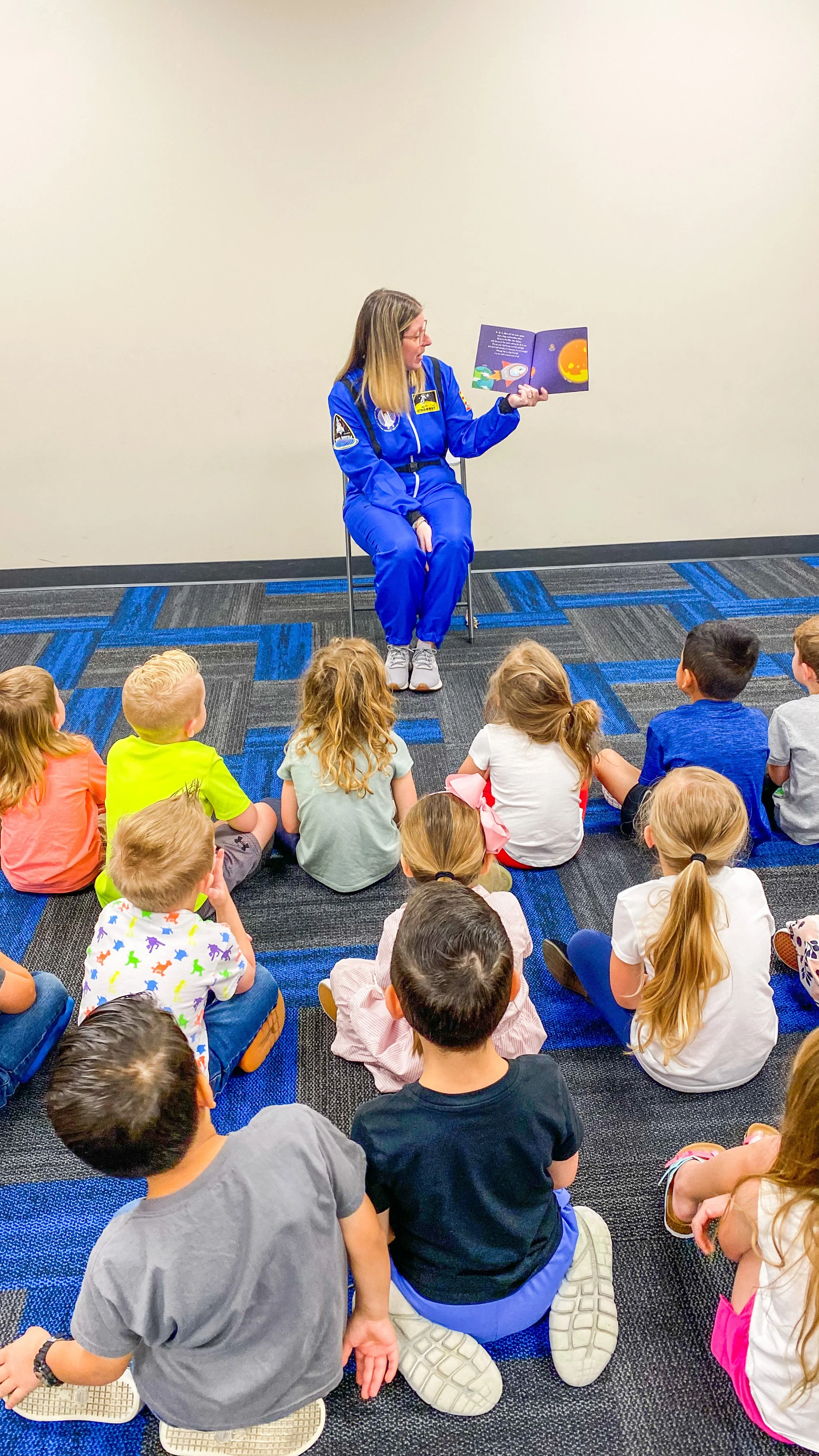 Amanda, the author of the Rowe+Rinn Books, is in an astronaut suit reading "Rowe+Rinn Blast into Space" to a group of young children seated on the floor, in a classroom setting.