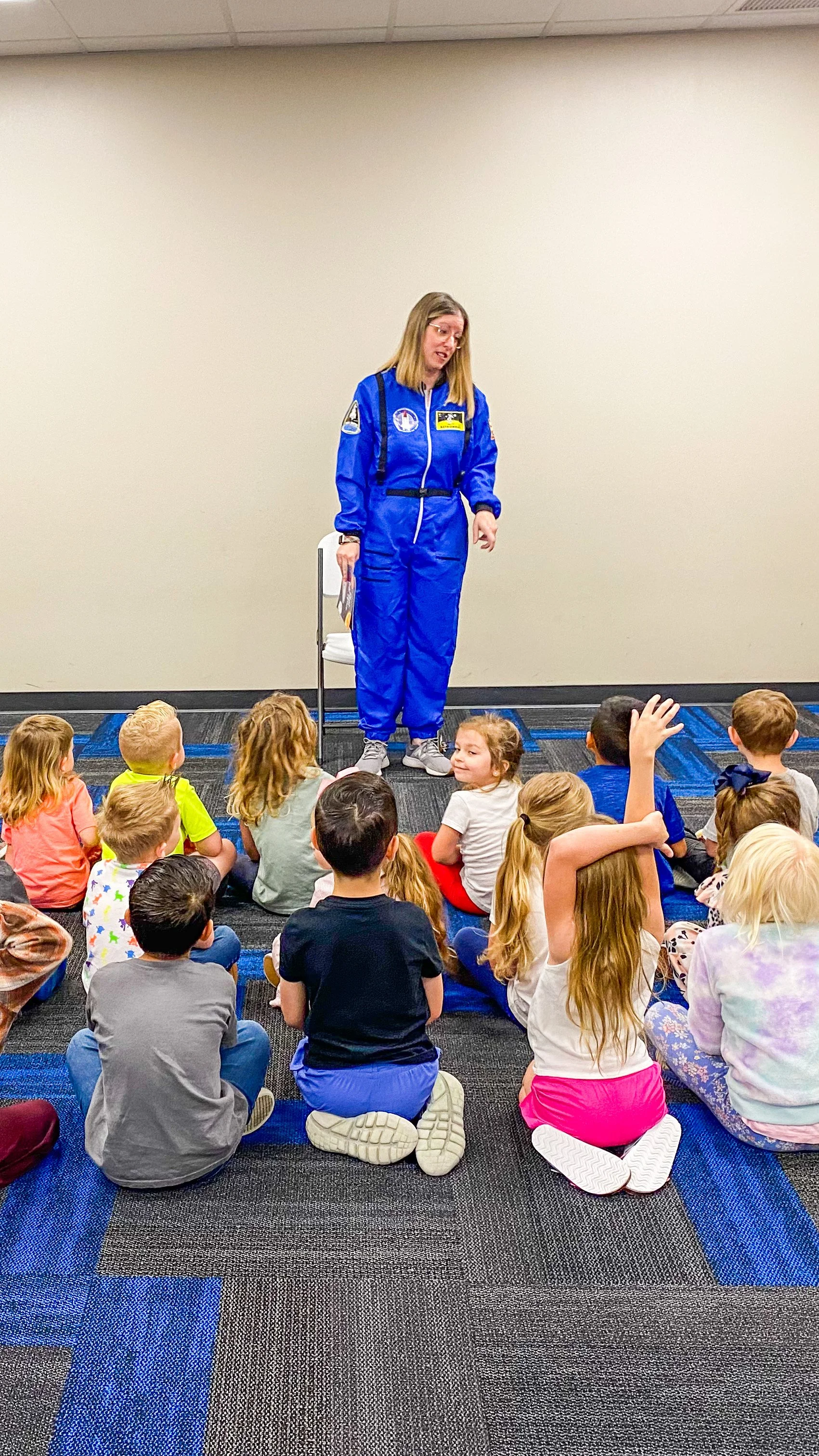 Amanda, the author of the Rowe+Rinn Books, is in a blue astronaut suit stands in front of a group of seated children on a carpeted floor. She is talking and presenting to the children who are attentively listening, with one child raising a hand.