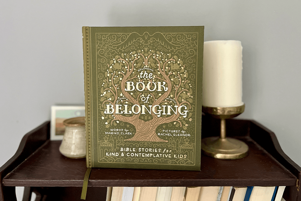 A hardcover children's book titled 'The Book of Belonging' by Mariko Clark with illustrations by Rachel Eleanor, placed on a wooden shelf, surrounded by a white candle, a small jar, and some framed photos.