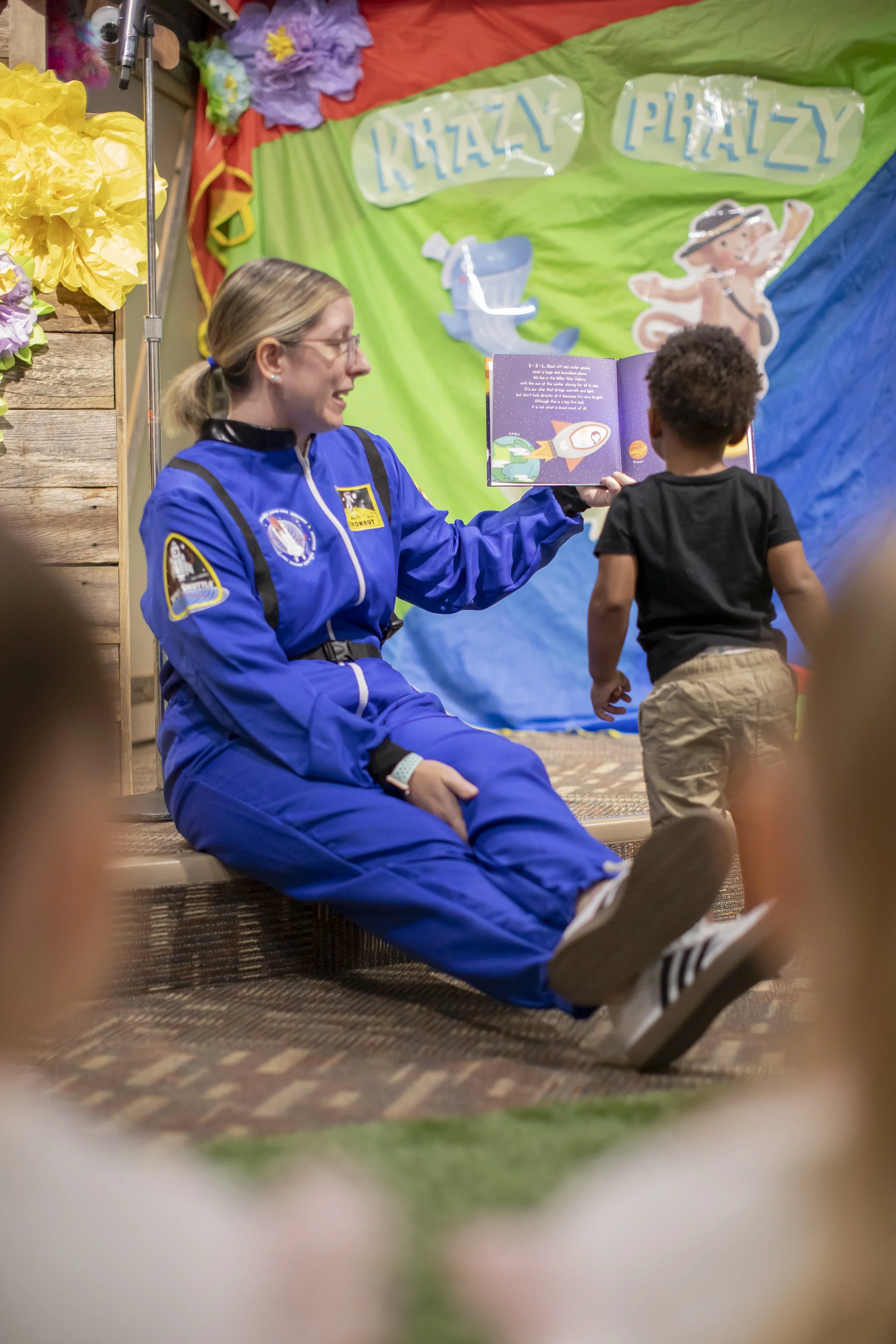Amanda, the author of the Rowe+Rinn Books, is in an astronaut costume reading to children at an event with colorful decorations. 