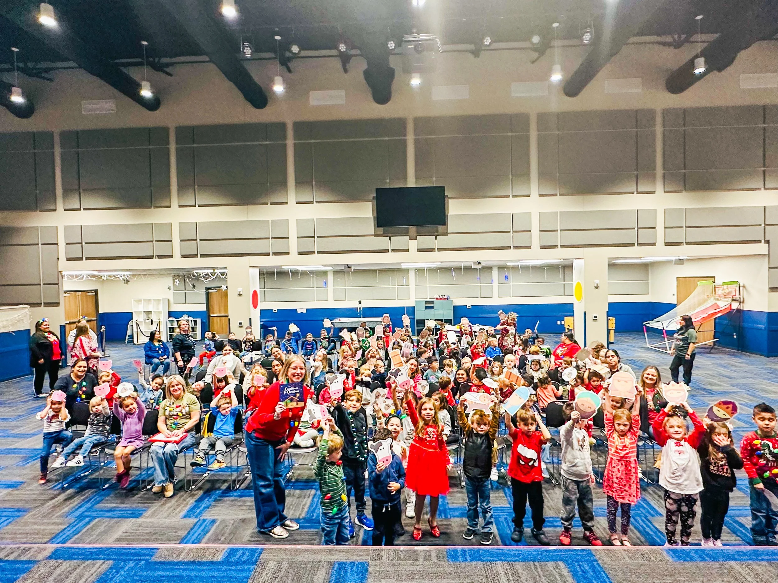 A large group of children and adults in a brightly lit auditorium or gym, some holding up colorful cut-out masks. The children are seated and dressed in casual clothing, with a few adults standing near them. The room has a blue and gray theme with hi