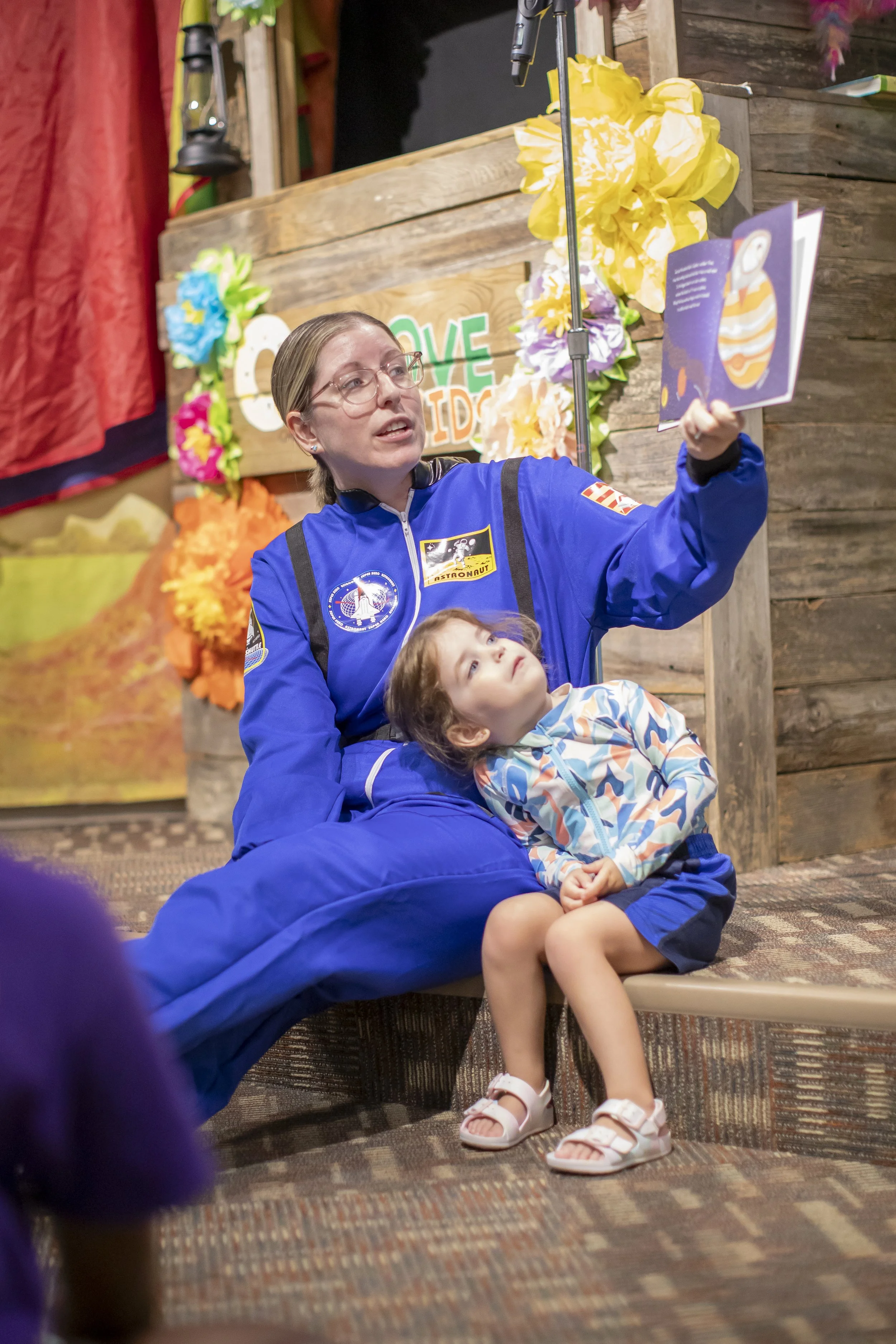 Amanda, the author of the Rowe+Rinn Books ,is wearing a blue astronaut suit reading a book to a young children with one on her lap, surrounded by colorful decorations.