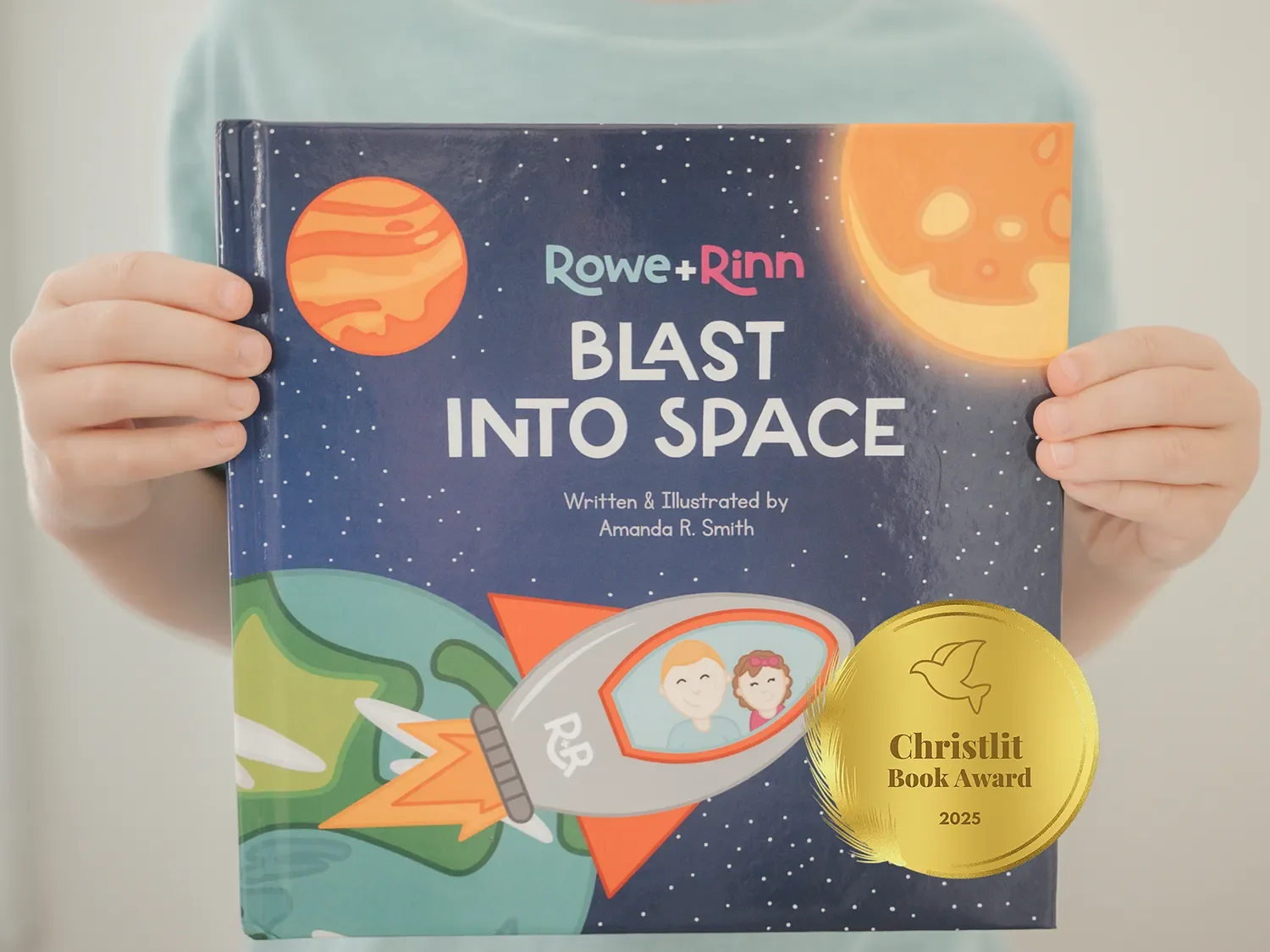 Child holding a children's book titled "Blast Into Space" by Amanda R. Smith, featuring colorful illustrations of a spaceship and planets.
