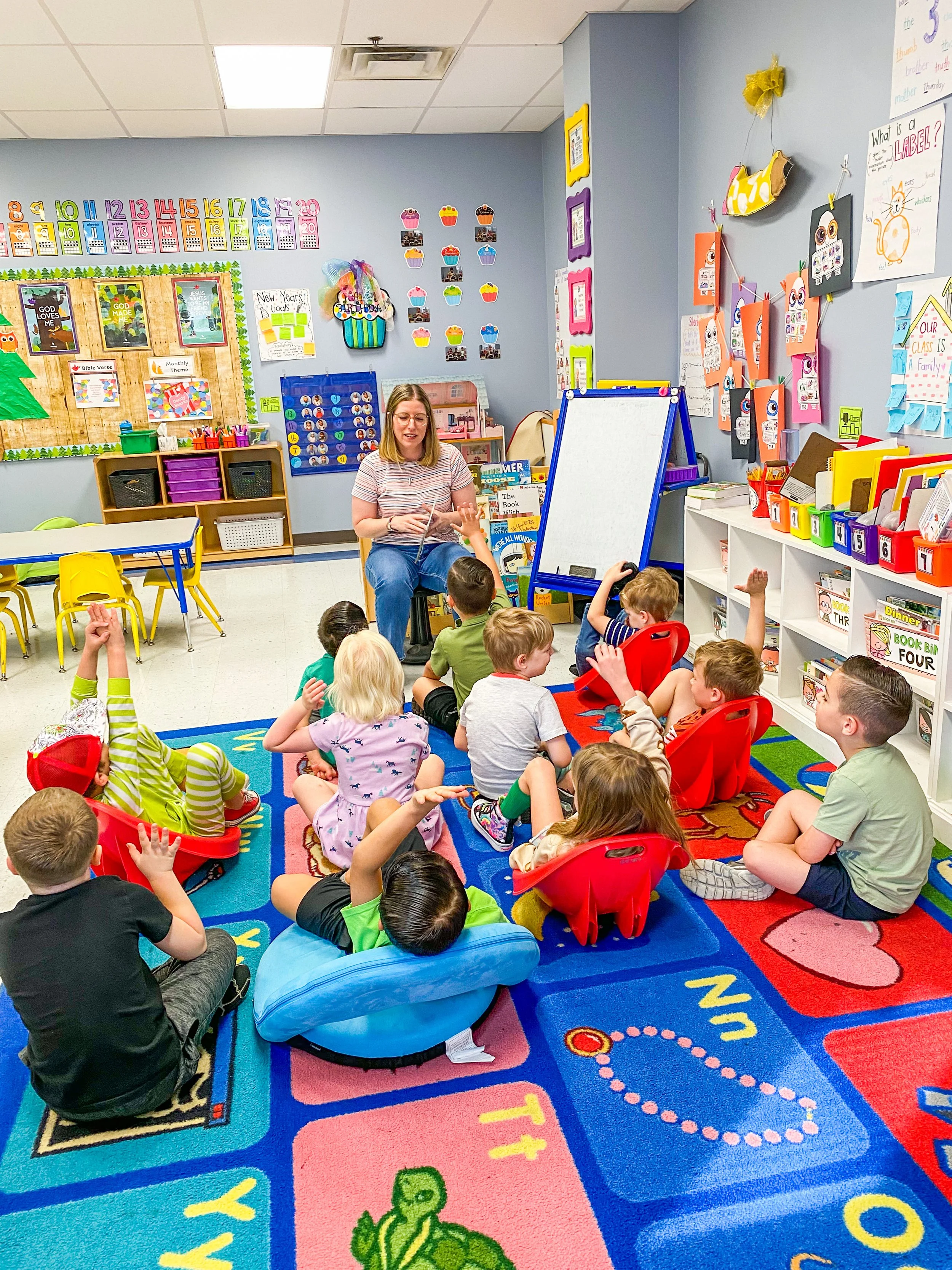 A teacher sits on a chair in a colorful elementary classroom filled with young children. The kids are seated on an alphabet-themed carpet, facing the teacher, with some raising their hands. The room features colorful educational decorations, includin
