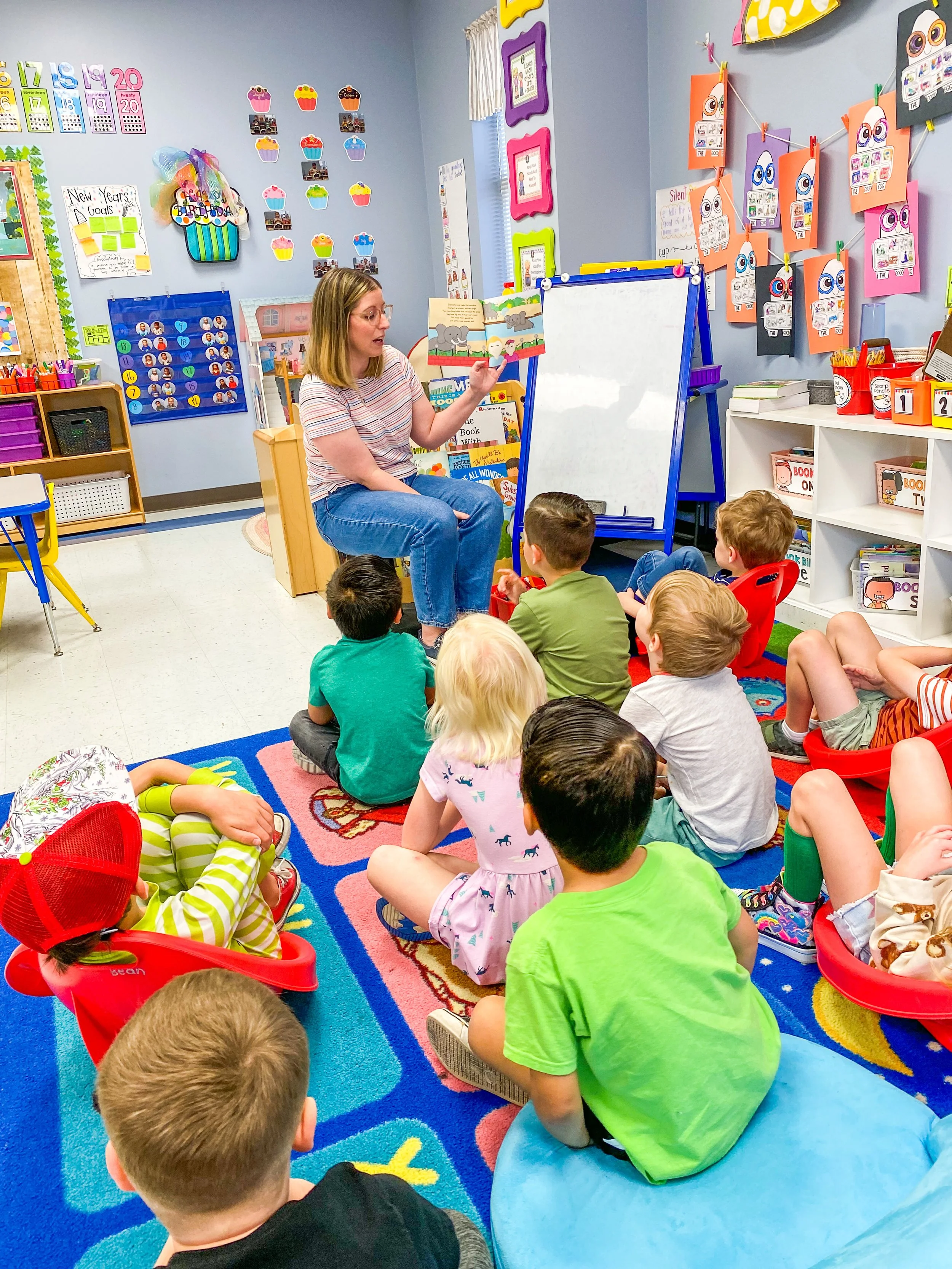 Amanda, the author of the Rowe+Rinn Books, is reading a book to a group of young children sitting on a colorful rug in a classroom with educational decorations and materials.