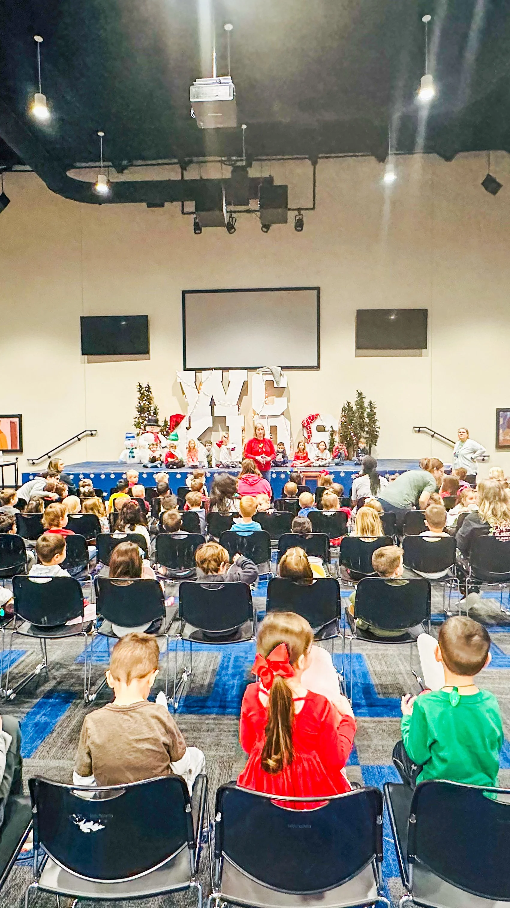 A group of children seated in an auditorium facing a stage with Christmas decorations and a speaker addressing them. The stage features festive decor including large white letters and a Christmas tree. The audience includes children and adults, some 