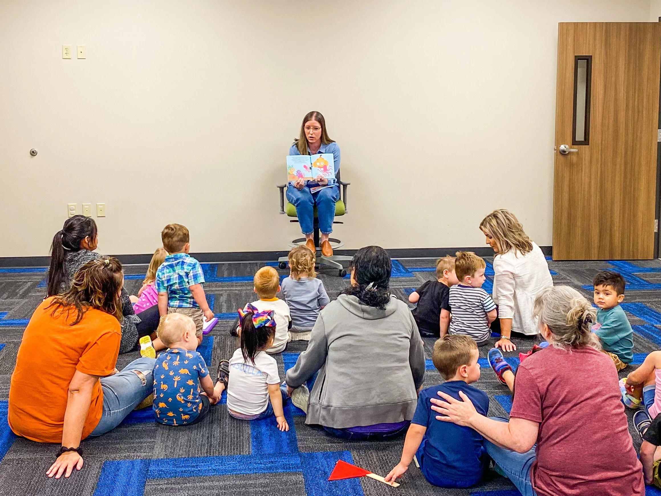 Amanda, the author of the Rowe+Rinn Books, is reading a picture book to a group of young children and adults seated on a carpeted floor in a room with a closed door.
