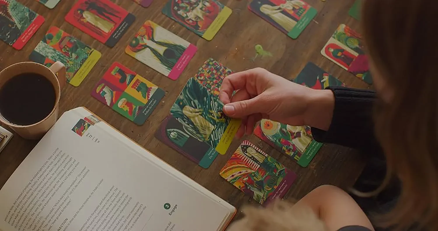 A person sitting at a wooden table, holding a colorful tarot card, with several other tarot cards laid out on the table. There is an open book with text, and a cup of coffee nearby.