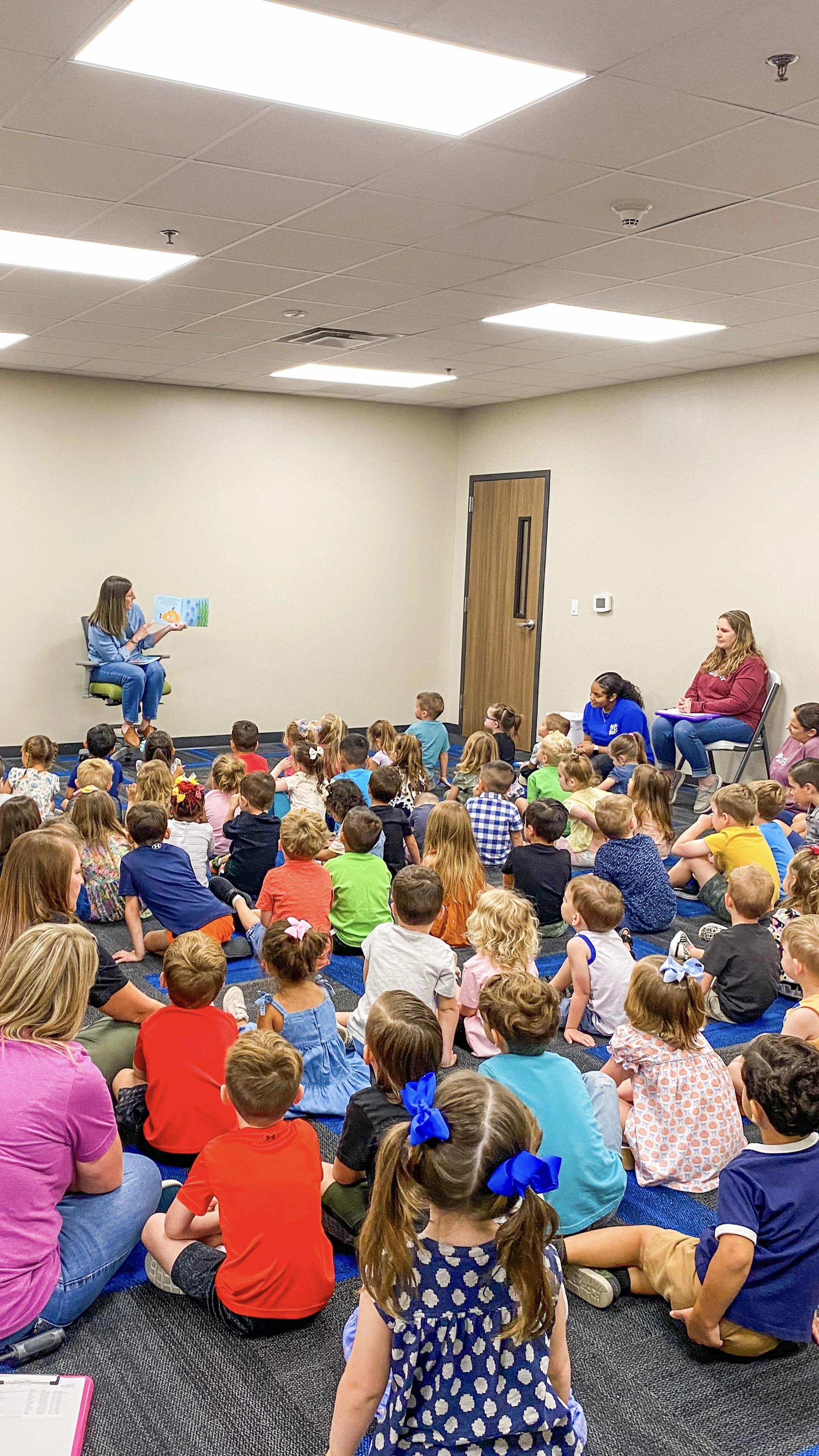 Amanda, the author of the Rowe+Rinn Books, is reading a picture book to a large group of young children sitting on the floor in a classroom setting. Two women are seated on chairs observing. The room has neutral walls and bright overhead lighting.