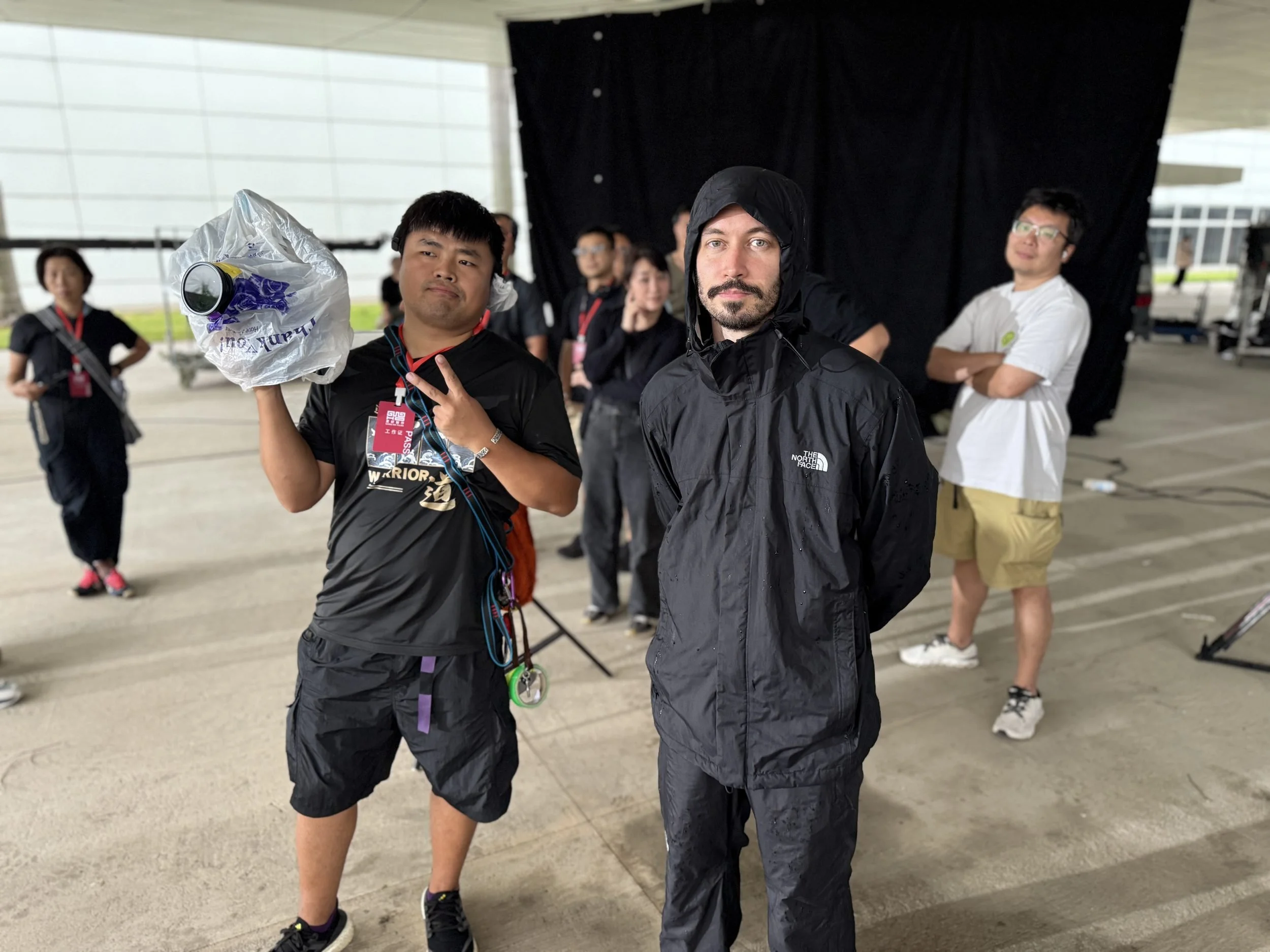 Zac waits in rain gear on a concrete set while a member of the local production crew holds a wrapped camera, flashing a peace sign.