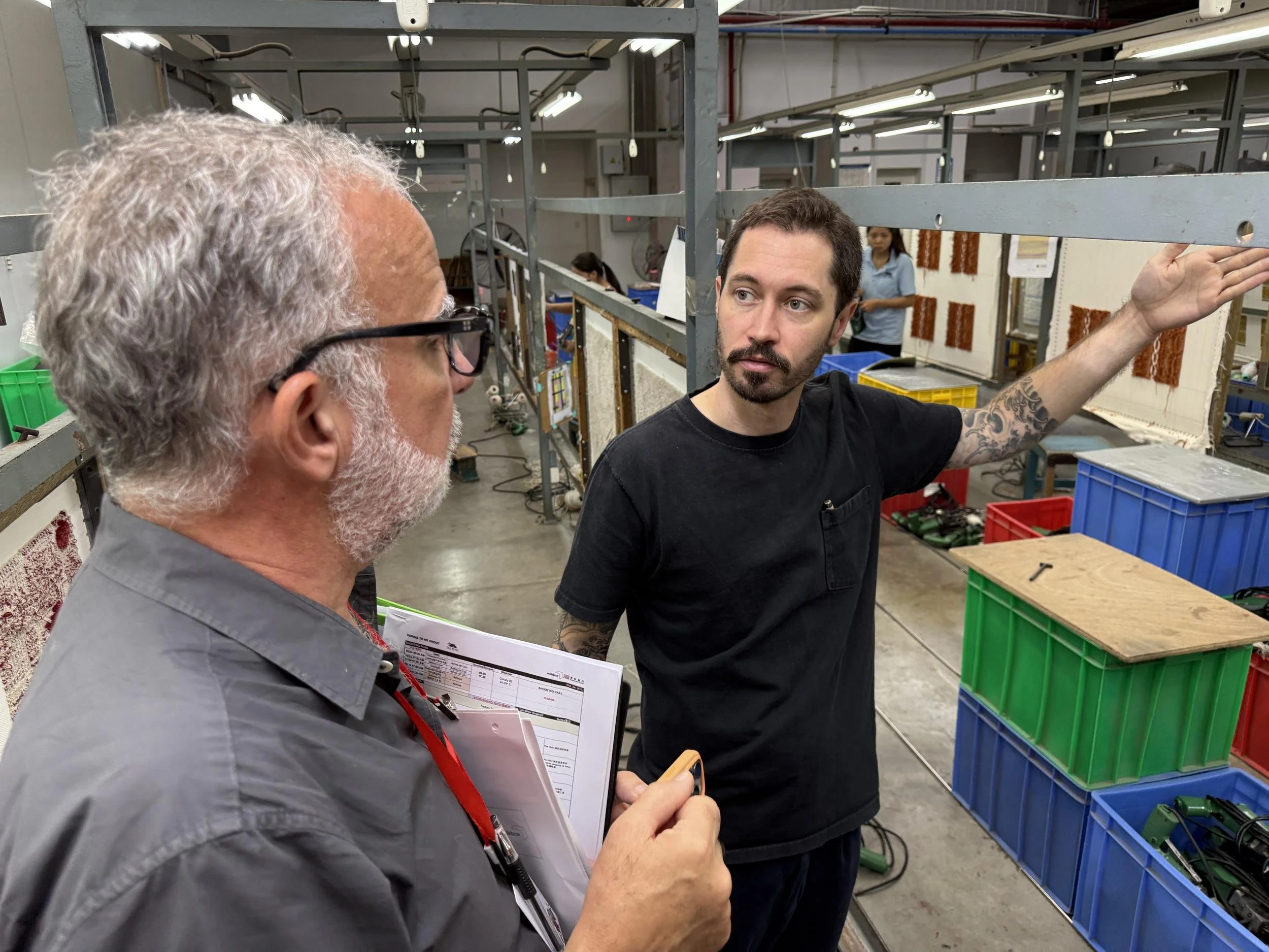 Zac and Ed talk on a factory floor, while Zac gestures toward racks of samples and stacked bins.