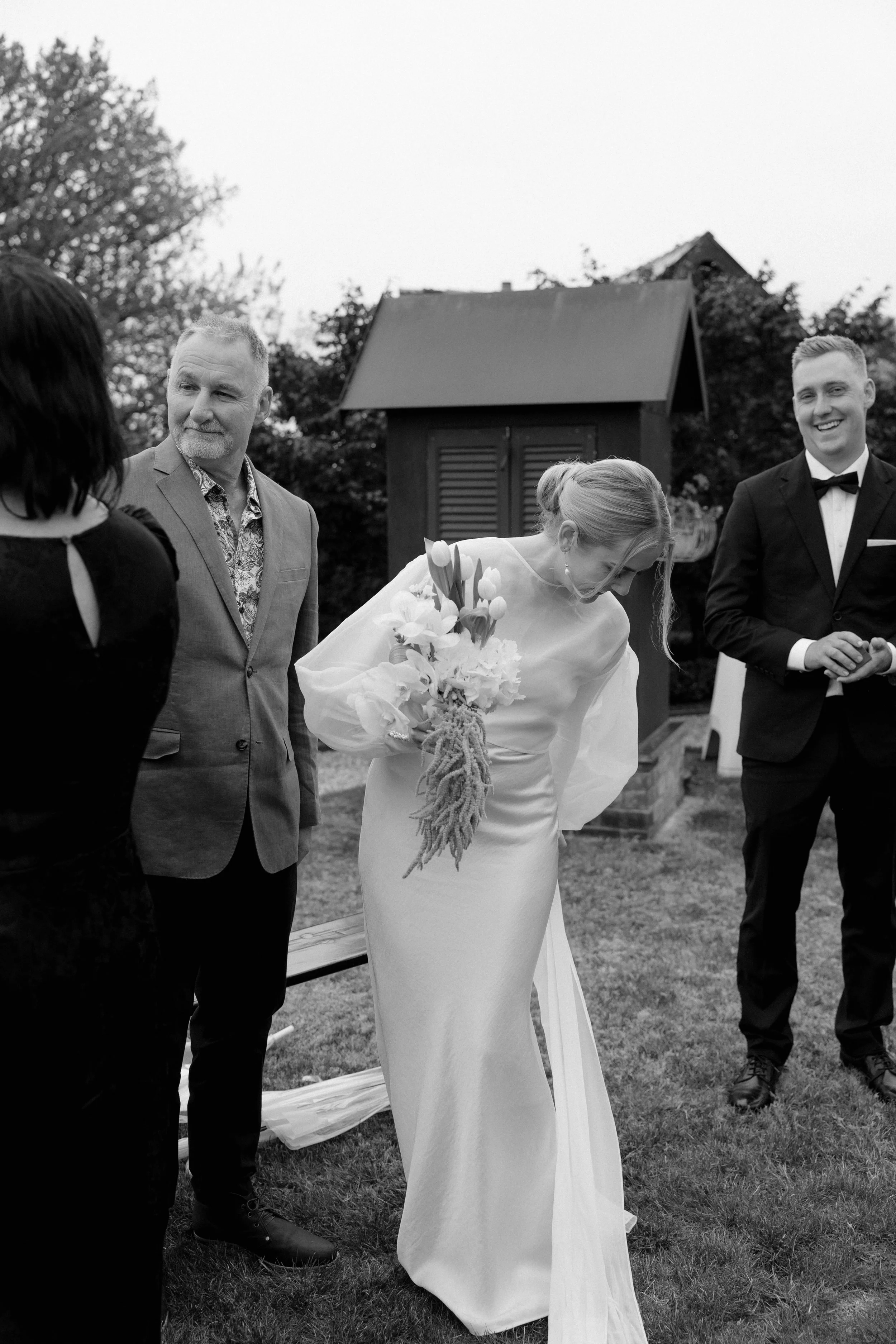 Black and white photo of a wedding scene outdoors with five people, including a bride in a white gown holding a bouquet, and four men in suits, two of whom are wearing tuxedos, standing on grass near a small wooden building with a sloped roof and tre