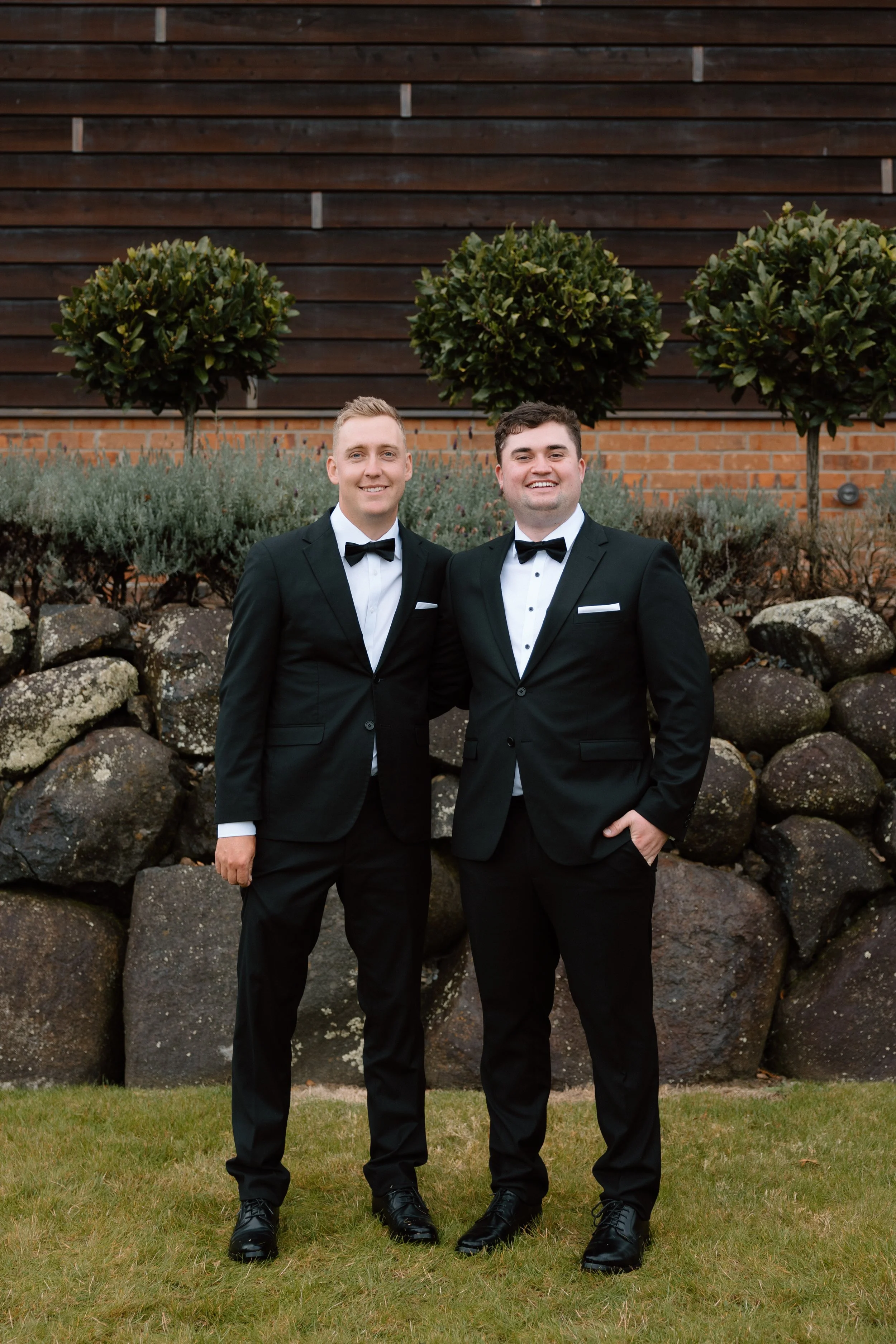 Two men in black tuxedos with bow ties standing outdoors, smiling, in front of a wooden fence and trimmed bushes, on a grassy area.