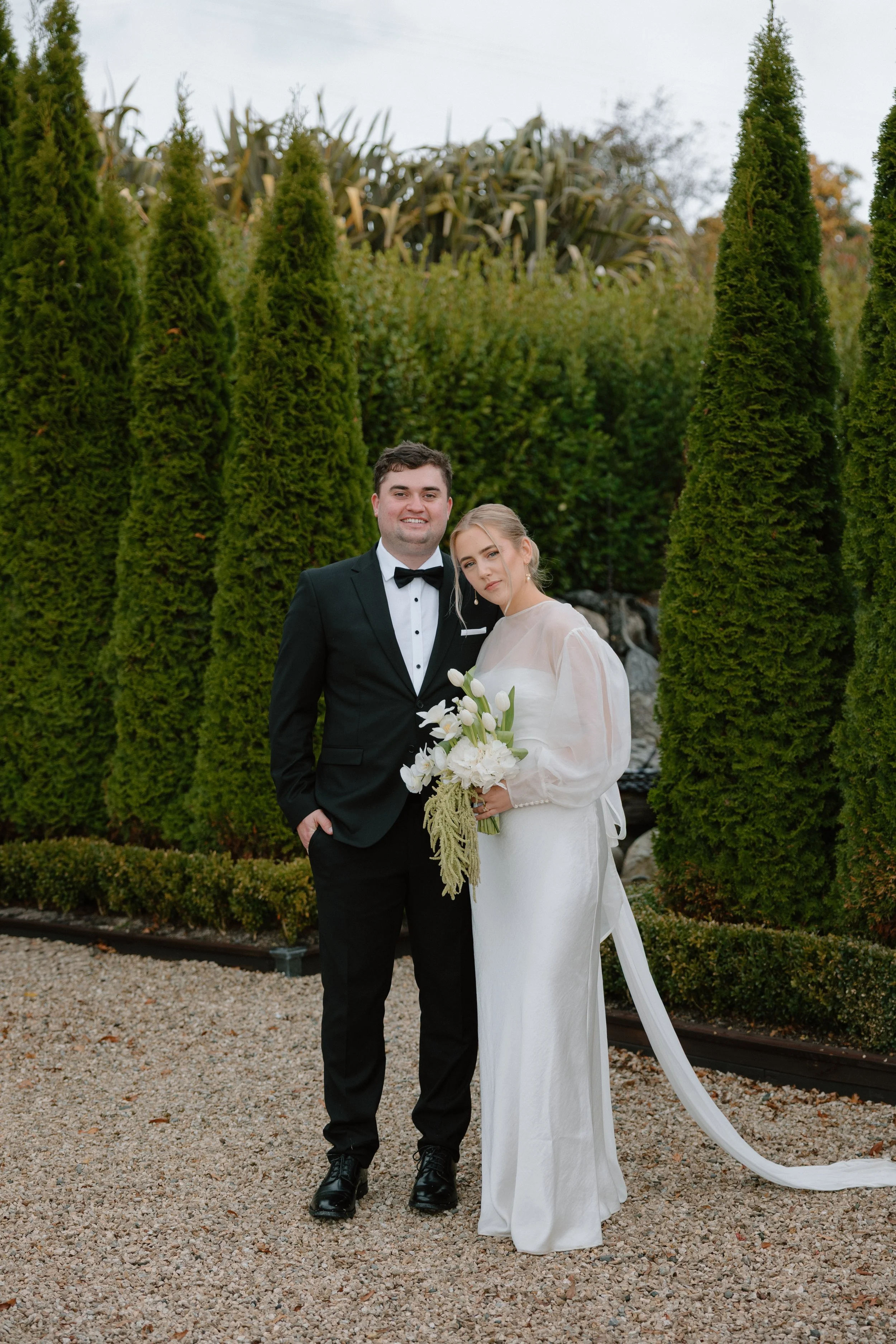 A newlywed couple stands outdoors among tall, green, coniferous trees. The groom wears a black tuxedo with a white shirt and black bow tie, smiling confidently. The bride wears a white wedding gown with sheer long sleeves, holding a bouquet of white 