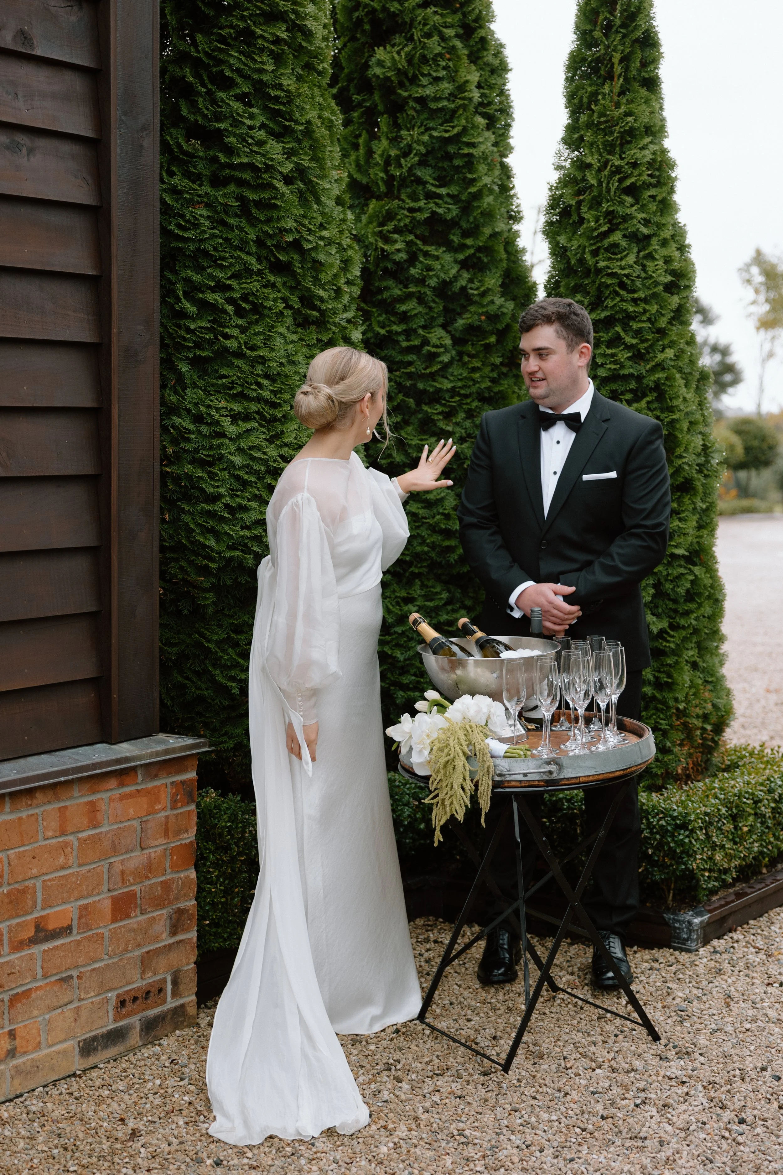 A woman and a man in tuxedo talking outdoors next to a tall hedge. The woman is wearing a white dress and has her hair in a bun. The man is wearing a black tuxedo with a bow tie. There is a small table with champagne bottles and glasses in front of t