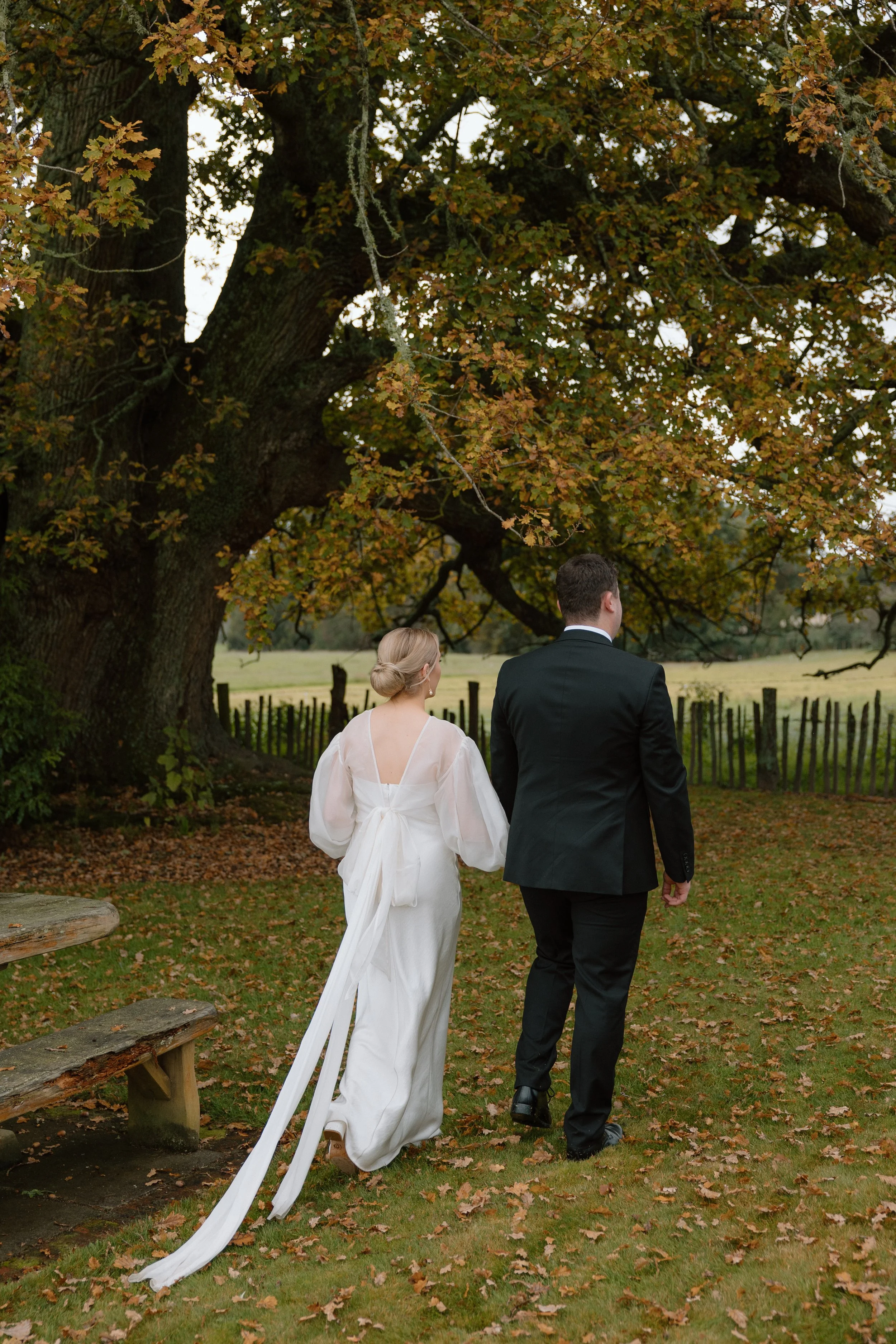 A bride and groom walking outdoors under a large, sprawling tree with fall leaves. The bride is in a white wedding dress and the groom in a black suit. They are walking away from the camera, holding hands.