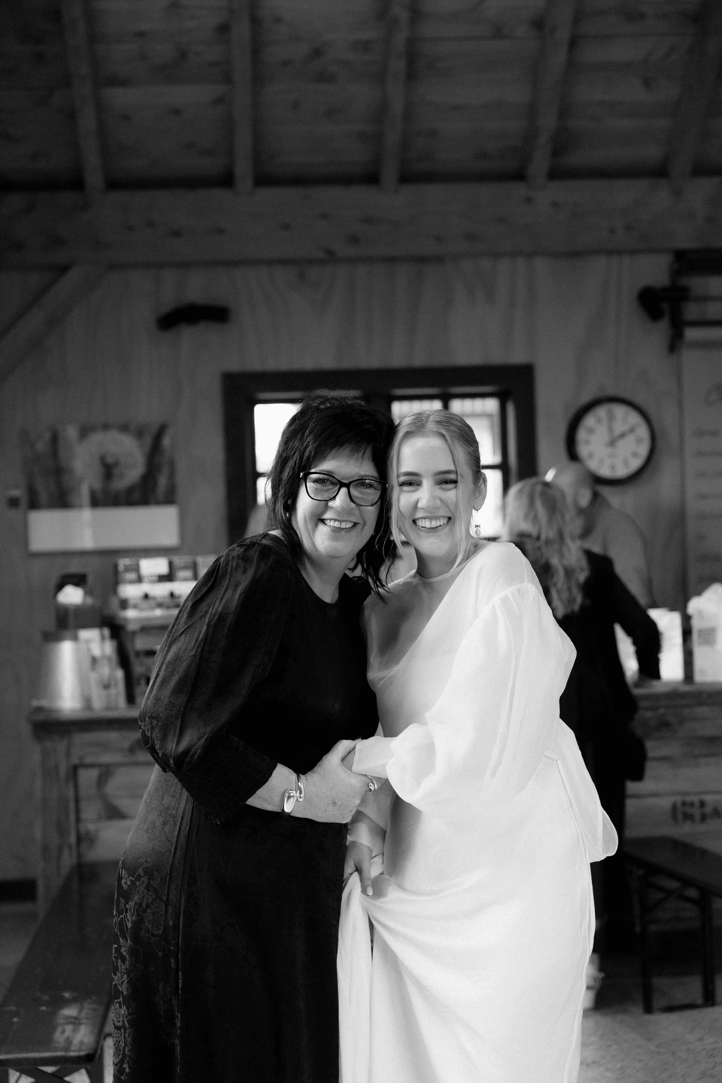 Two women smiling and embracing in a cozy room with wooden walls. One woman with glasses and dark hair wearing a dark dress, the other with blonde hair and earrings wearing a white dress. A clock and a window are visible in the background.