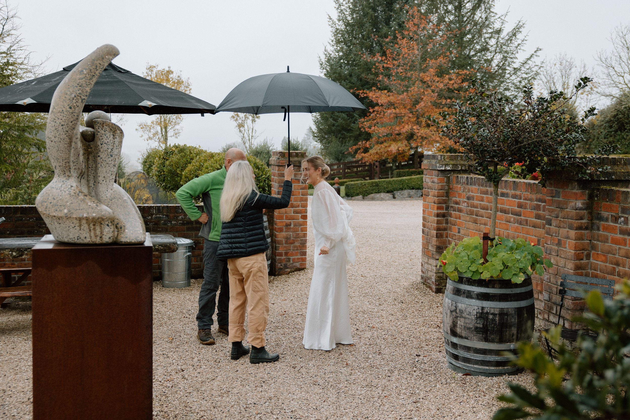 People gathered outdoors under umbrellas, with a woman in a white dress, possibly a bride, talking to two men and another woman, in a setting with brick walls, trees with autumn foliage, and decorative statues.