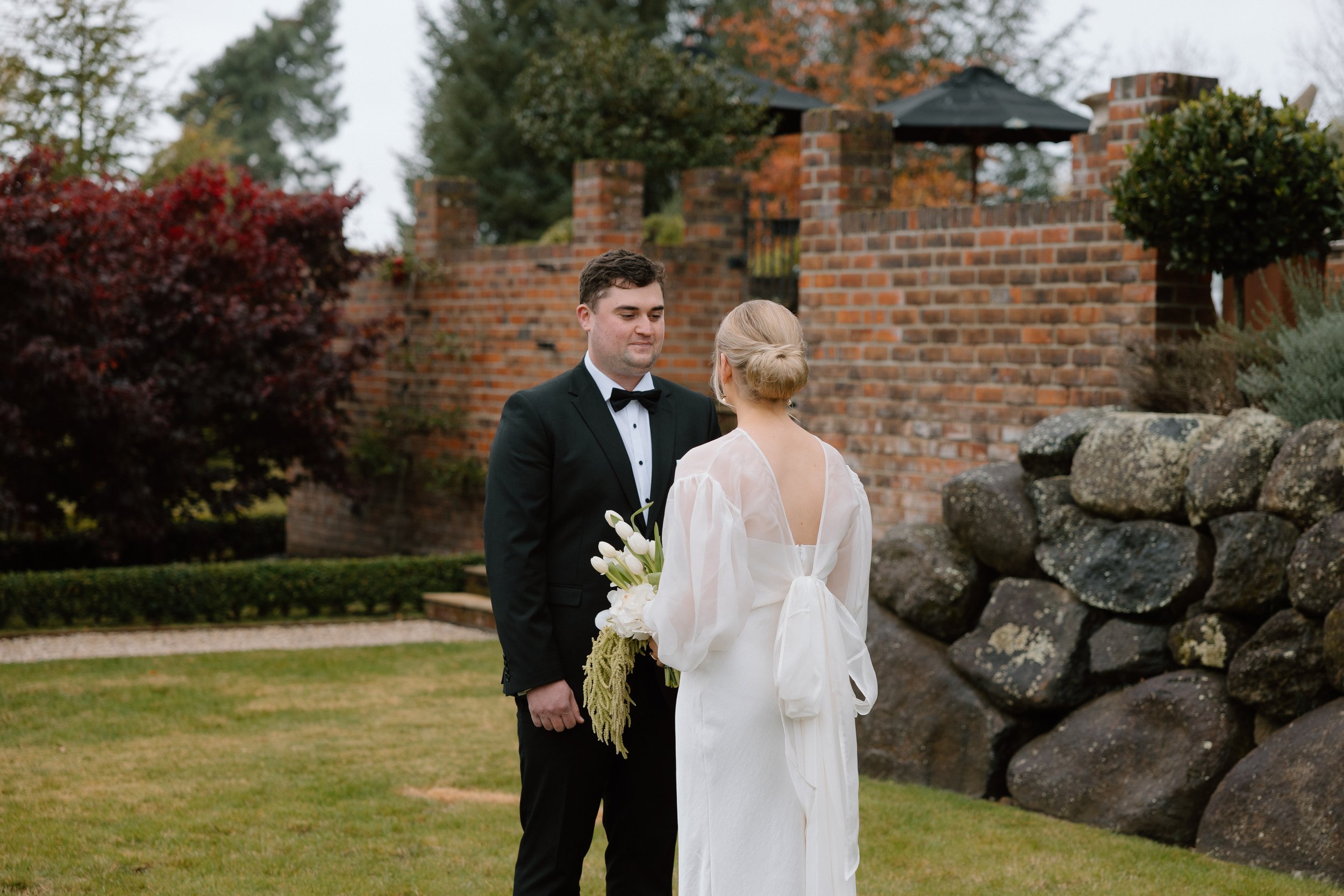 A bride and groom standing outdoors during a wedding ceremony. The groom is dressed in a black tuxedo with a bow tie, and the bride is wearing a white gown and holding a bouquet of white flowers. They are facing each other with a brick wall, trees, a