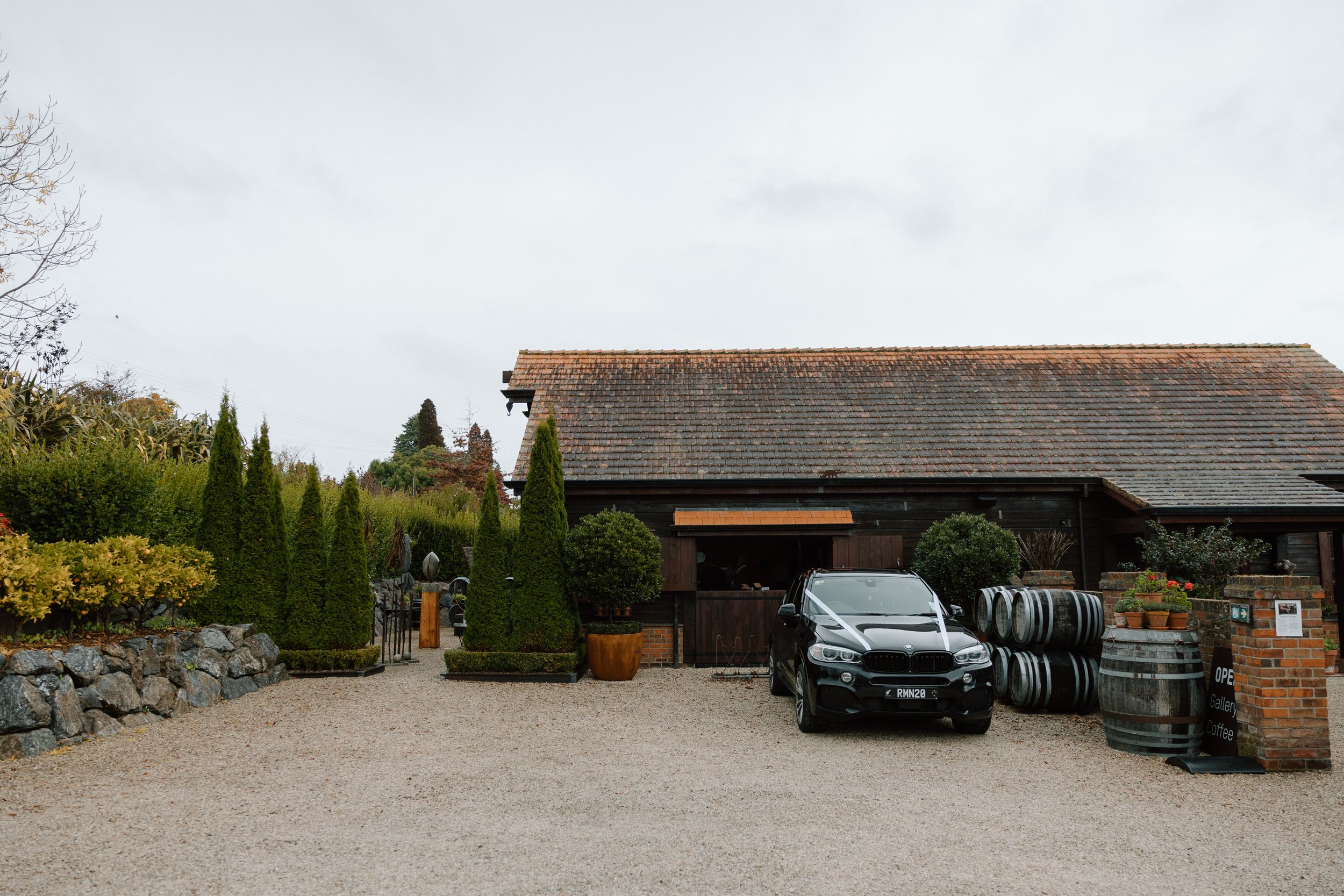 A gravel driveway in front of a rustic building with a tiled roof, surrounded by trees and potted plants, with cars parked near the entrance and barrels stacked on the right.