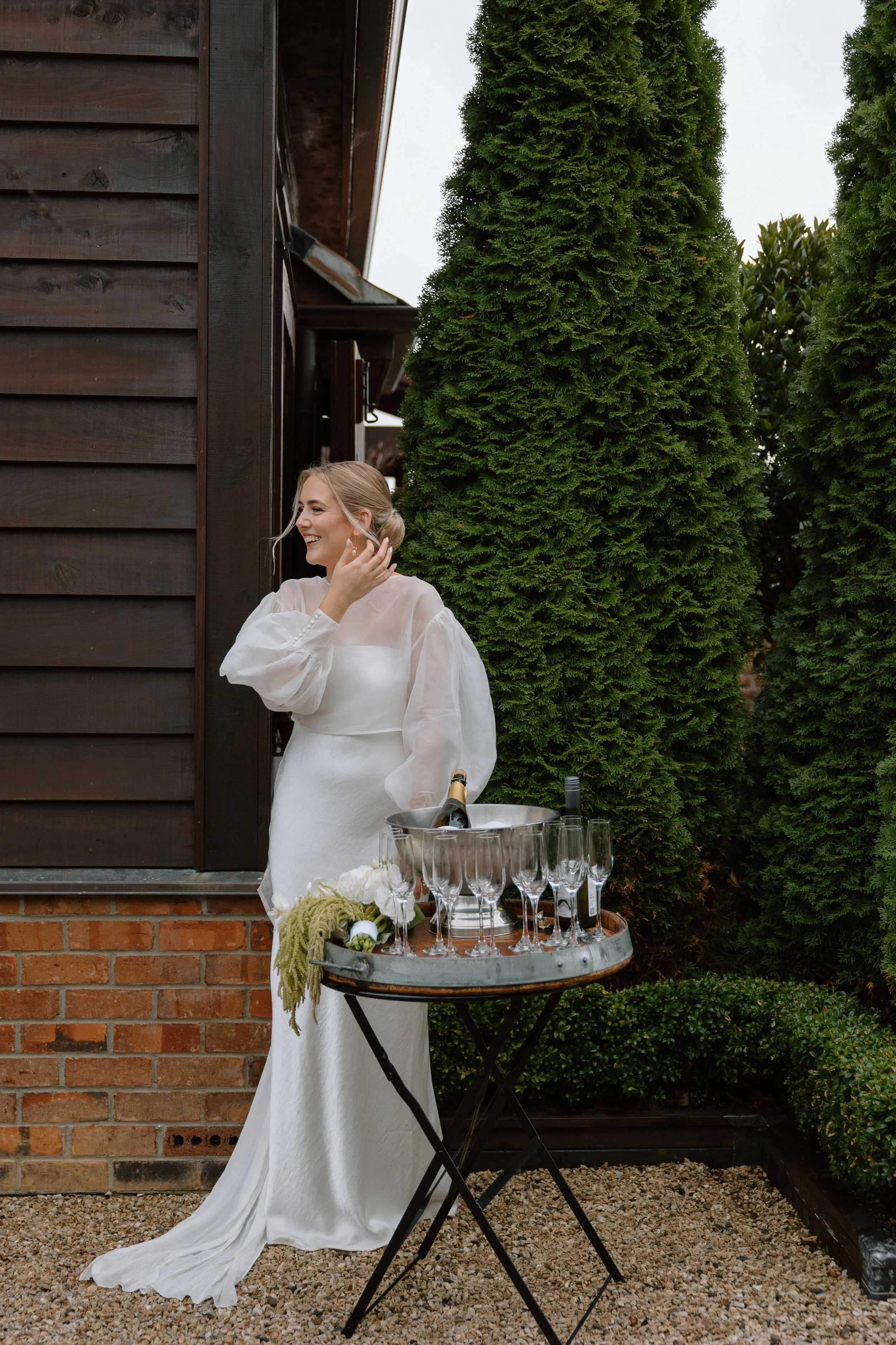 A woman in a white dress smiling beside a table with champagne glasses and a wine bottle outdoors