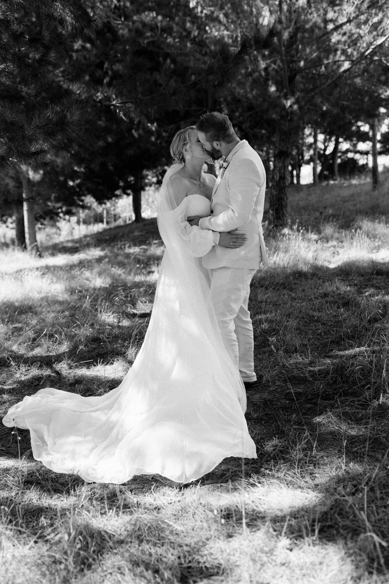 A black and white photograph of a bride and groom kissing outdoors among trees, with the bride wearing a strapless wedding gown and the groom in a light-colored suit.