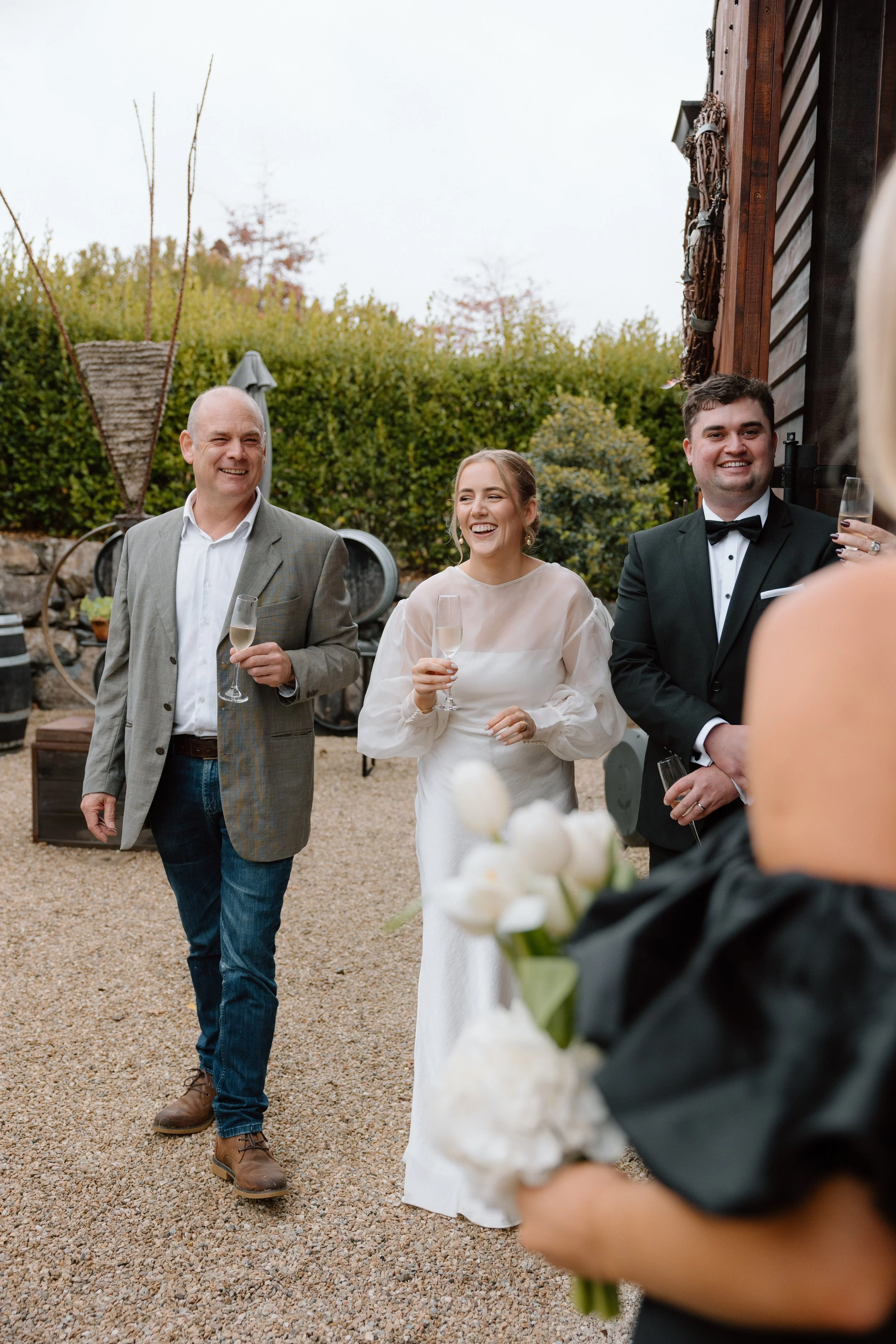 People at an outdoor wedding celebration, including a bride in a white wedding dress holding a glass of champagne, a man in a tuxedo with a bow tie also holding a glass of champagne, and an older man in a blazer with a green and brown plaid pattern h