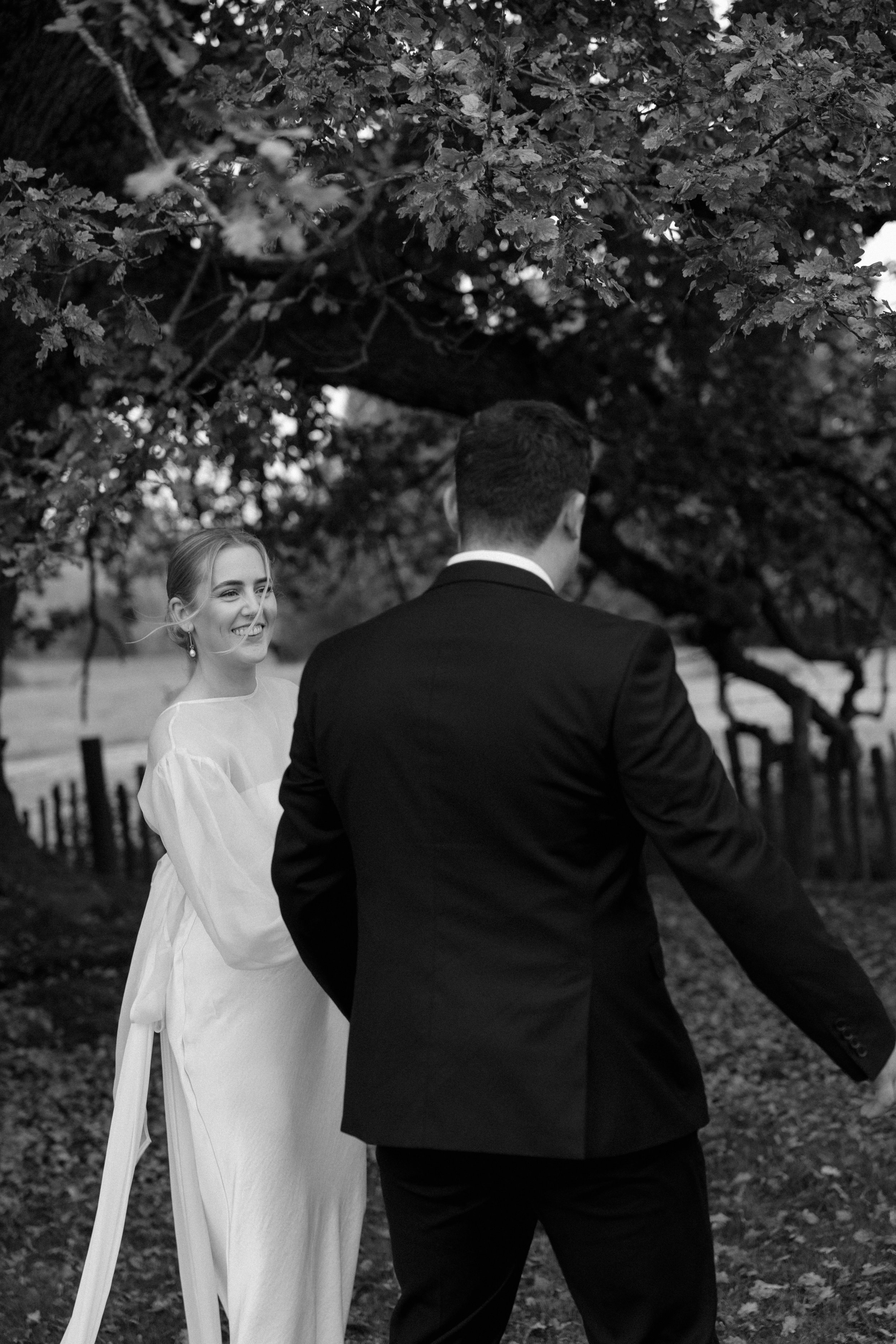 A black and white photo of a woman smiling and facing a man, outdoors under a large tree, with a fence and field in the background. The woman is wearing a light-colored dress or gown, while the man is in a suit.