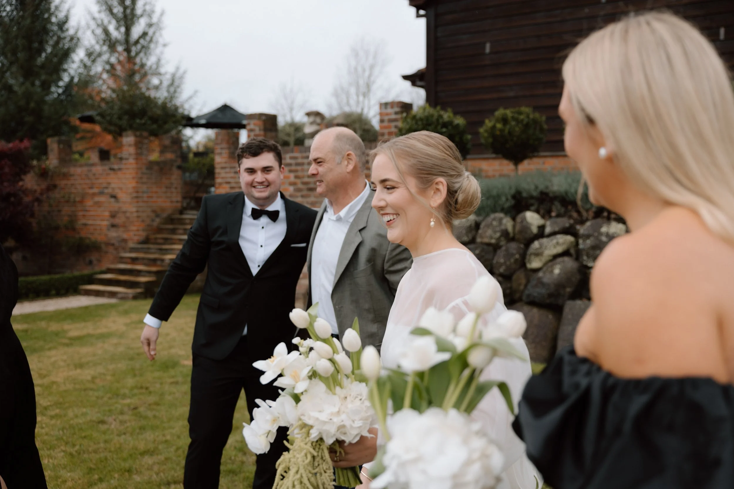 A group of people, including a woman in a white dress holding a bouquet of white flowers, walking outdoors on a cloudy day. A man in a tuxedo and another man in a gray suit are smiling and walking beside her. Another woman with blonde hair is partial