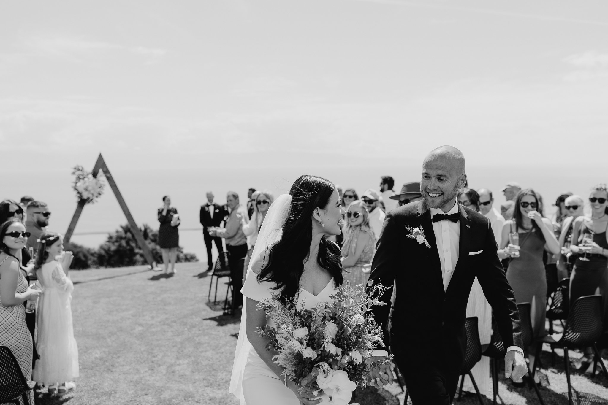 A bride and groom smiling and holding hands at their outdoor wedding ceremony, surrounded by guests in sunny weather.