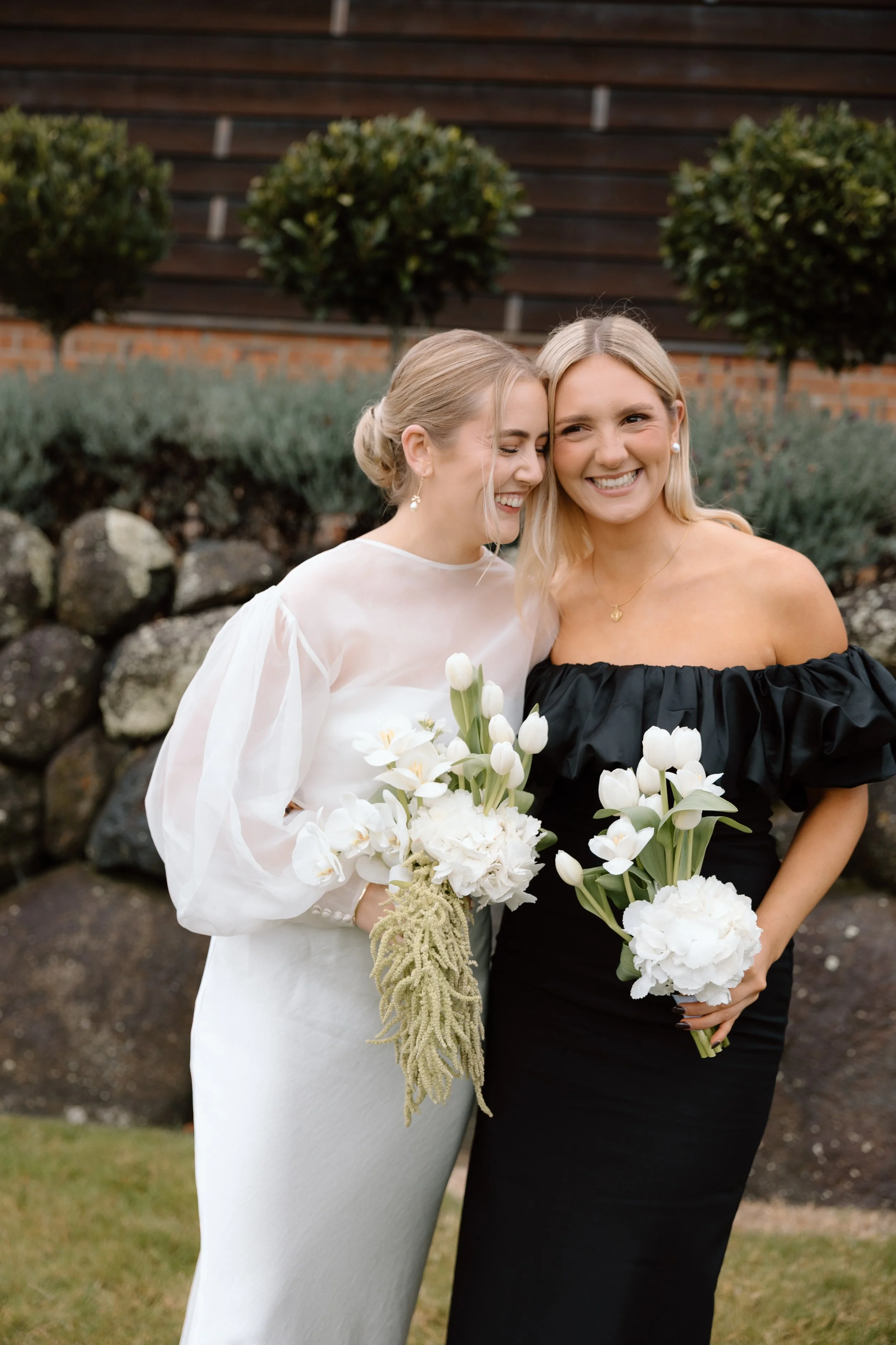Two women in elegant dresses, smiling and standing close together outdoors, holding bouquets of white flowers with a background of greenery and a brick wall.