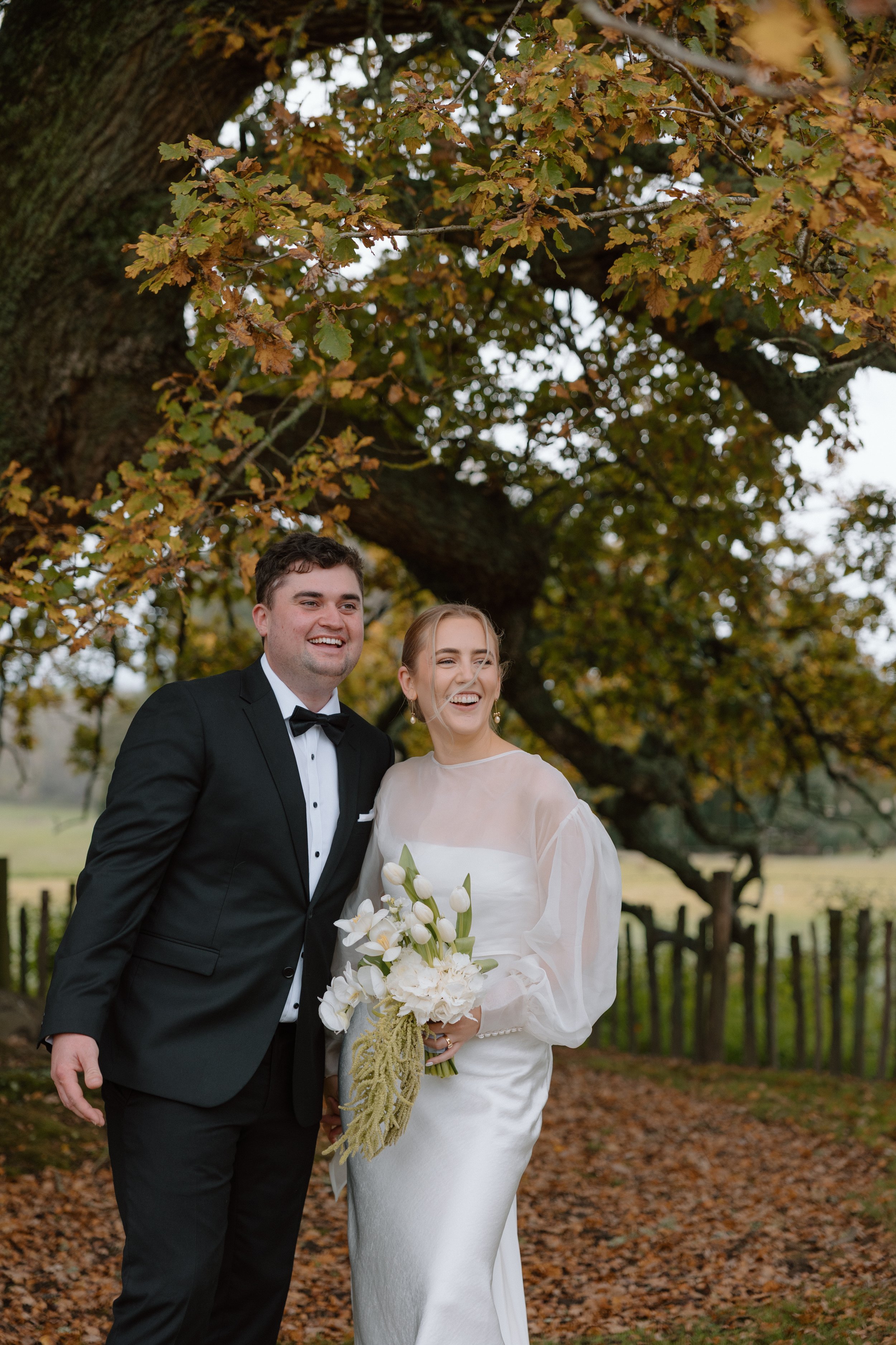 A bride and groom standing outdoors under a large tree with autumn-colored leaves, smiling happily during their wedding celebration. The groom is dressed in a black tuxedo and the bride wears a white wedding gown holding a bouquet of white flowers.