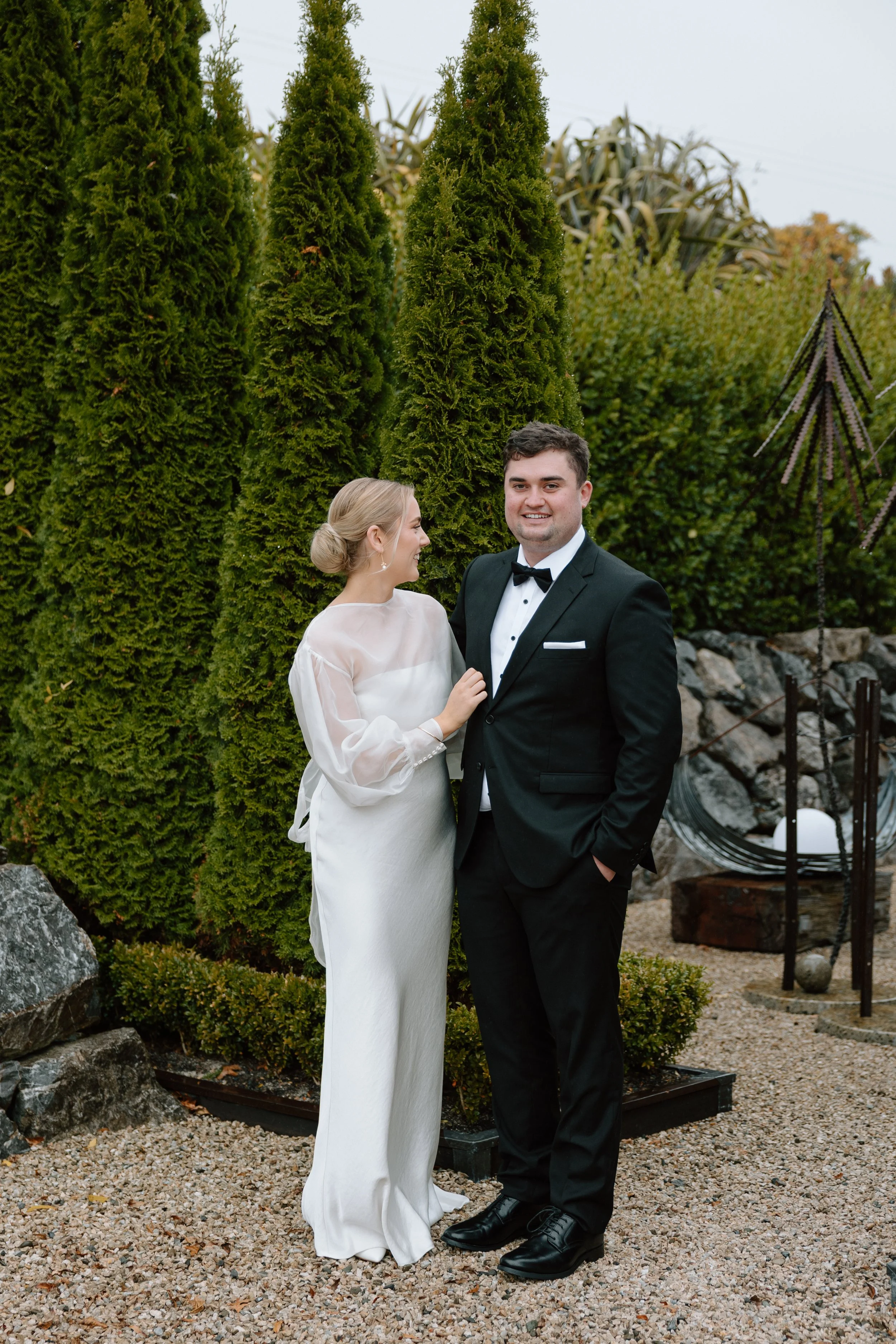 A bride and groom standing outdoors in front of tall evergreen trees and decorative plants. The bride is wearing a white gown and the groom is in a black suit with a bow tie, smiling at the camera.