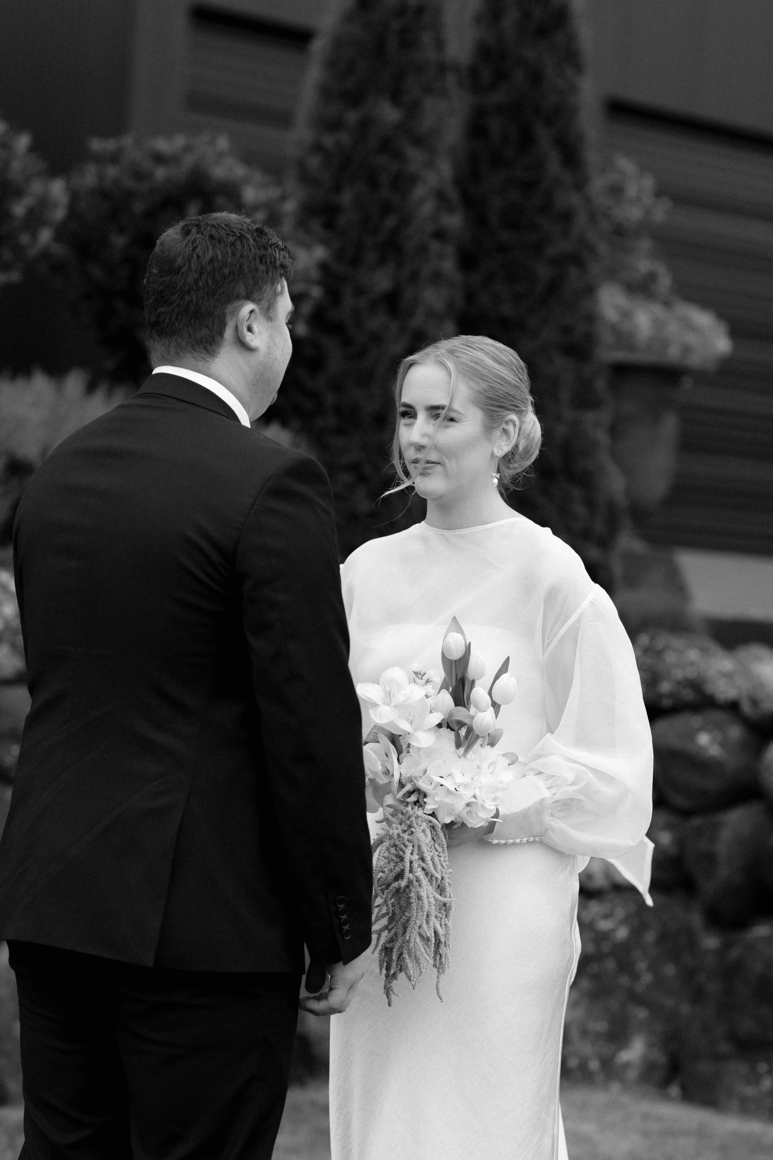 A bride and groom holding hands during a wedding ceremony outdoors, with the groom facing away and the bride looking at him, holding a bouquet of flowers.