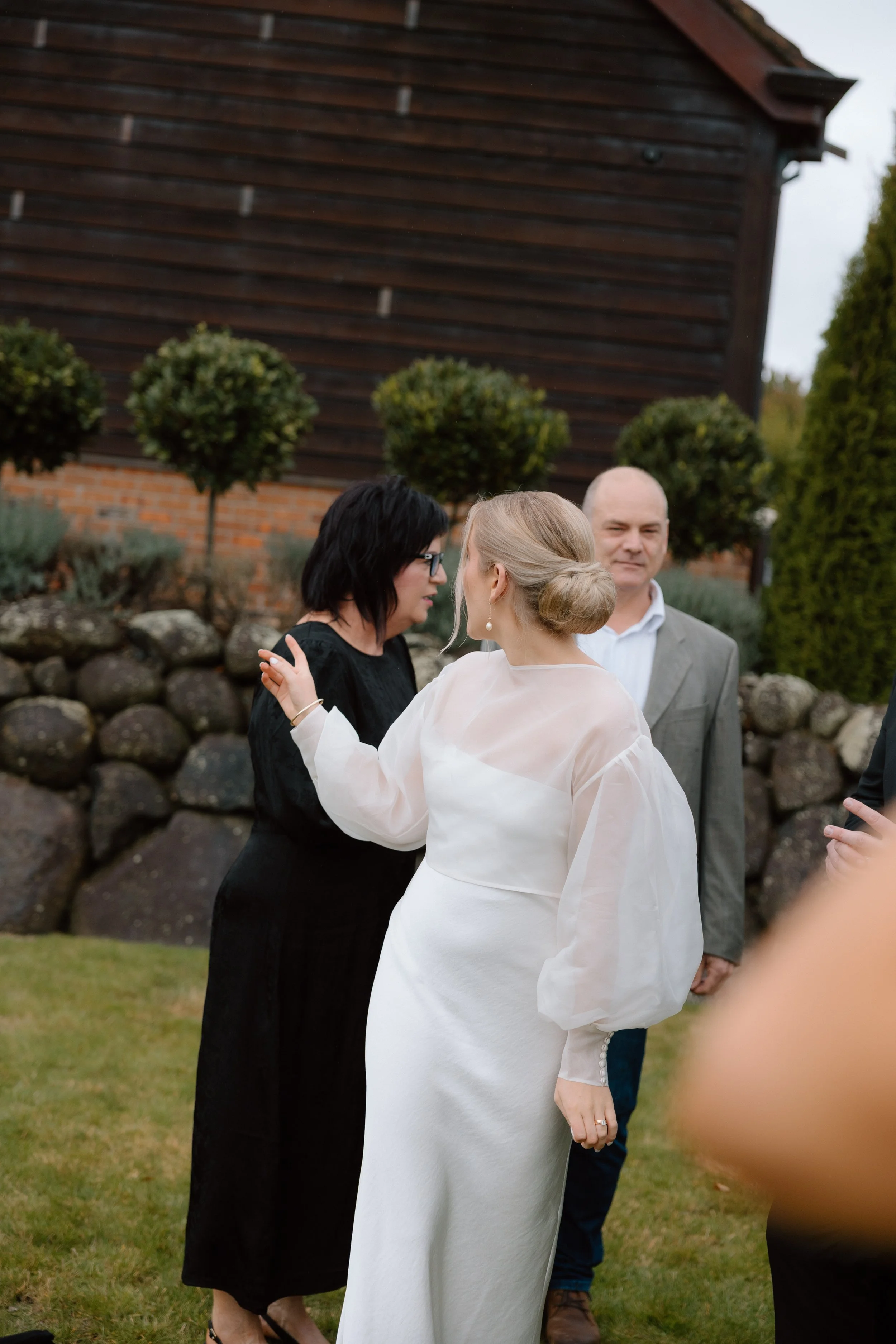 A woman in a white dress and pearl earrings is hugging and talking to another woman in a black dress during an outdoor gathering. A man in a gray suit stands behind them, smiling. The background features a stone wall, lush greenery, and a wooden buil