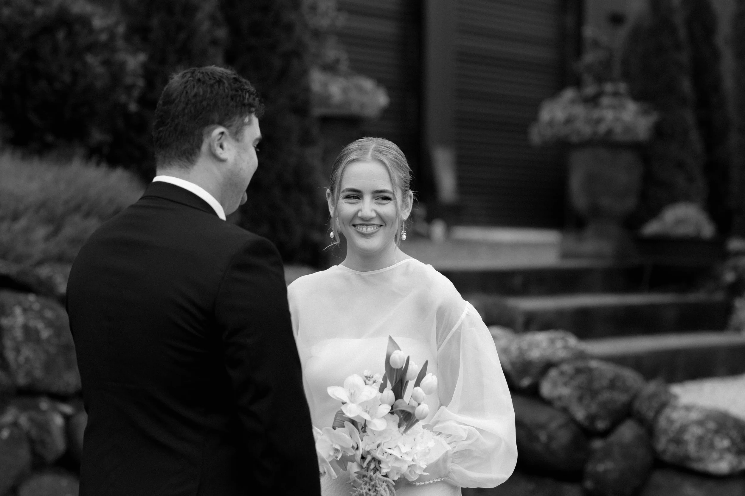 A black and white photo of a bride and groom standing outdoors facing each other, with the bride smiling and holding a bouquet of white flowers.
