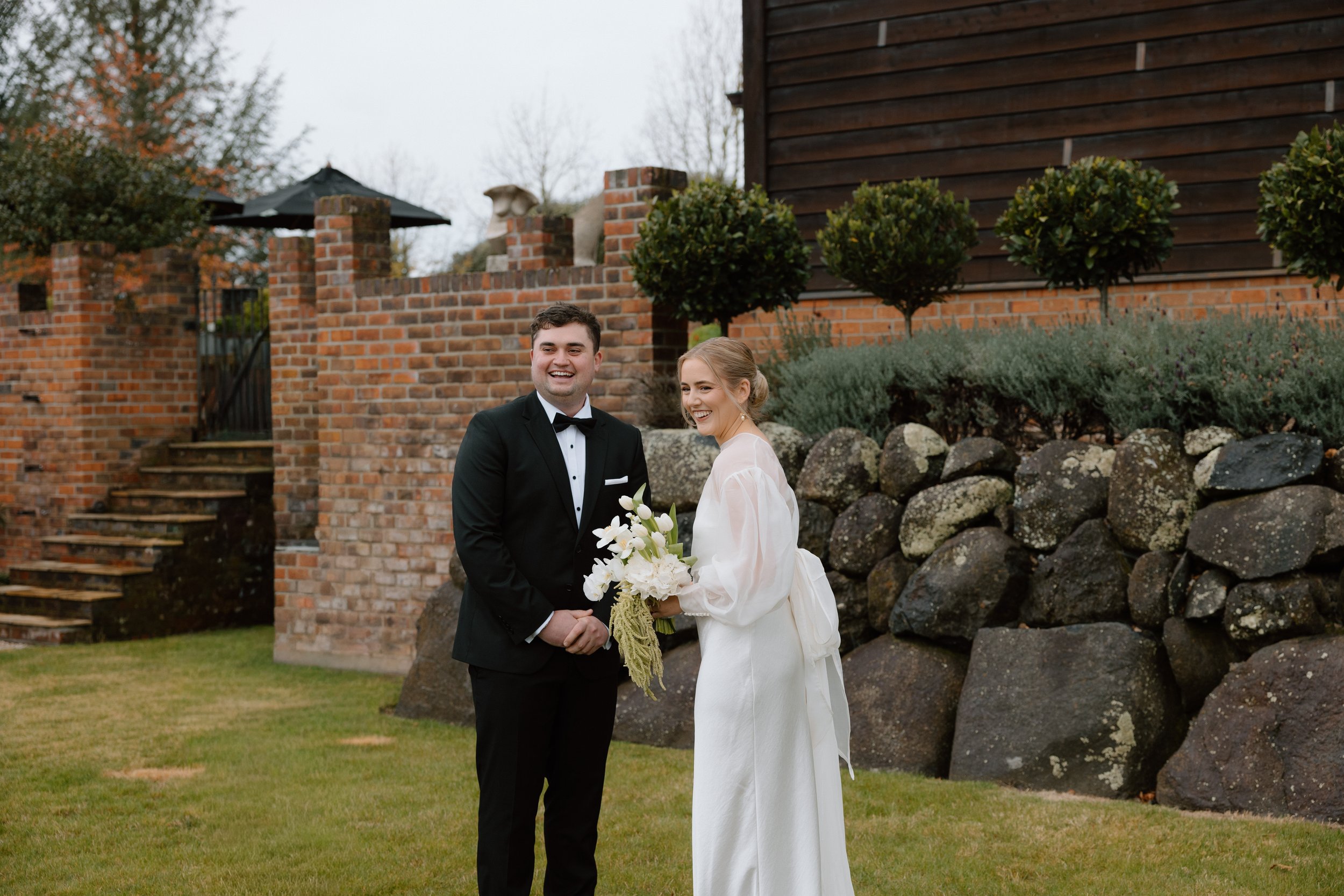 A newlywed couple holding hands and smiling outdoors in front of a brick and wooden fence with trees and bushes, during a cloudy day.