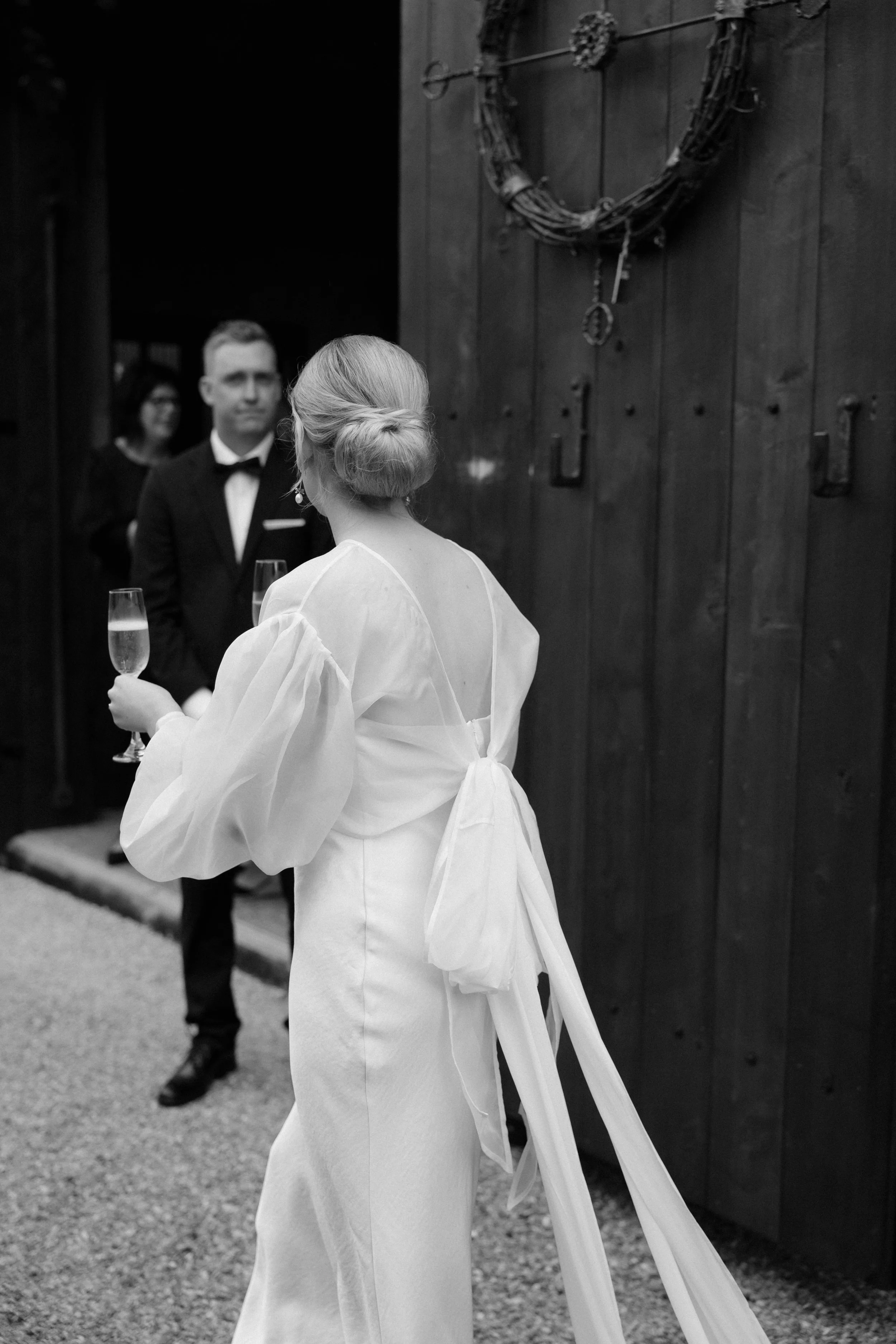 A woman in a white dress with her hair in a low bun holds a glass of champagne at a wedding or formal event, with a man in a tuxedo and bow tie and another person in the background, standing near a wooden wall decorated with a wreath.