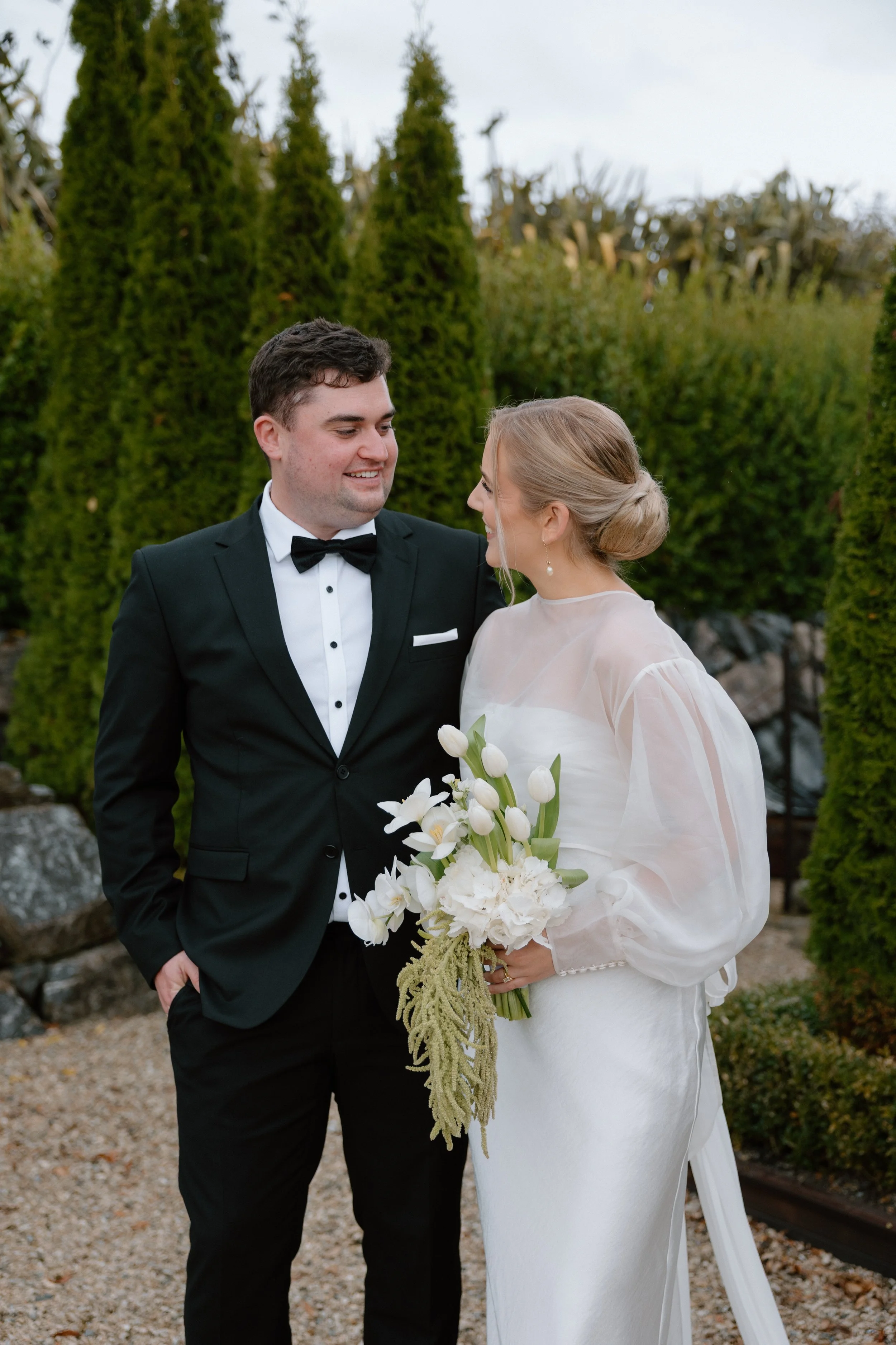 A newlywed couple dressed in formal wedding attire stands outside among greenery and rocks, smiling at each other. The groom wears a black tuxedo with a bow tie, and the bride wears a white wedding dress with sheer long sleeves, holding a bouquet of 