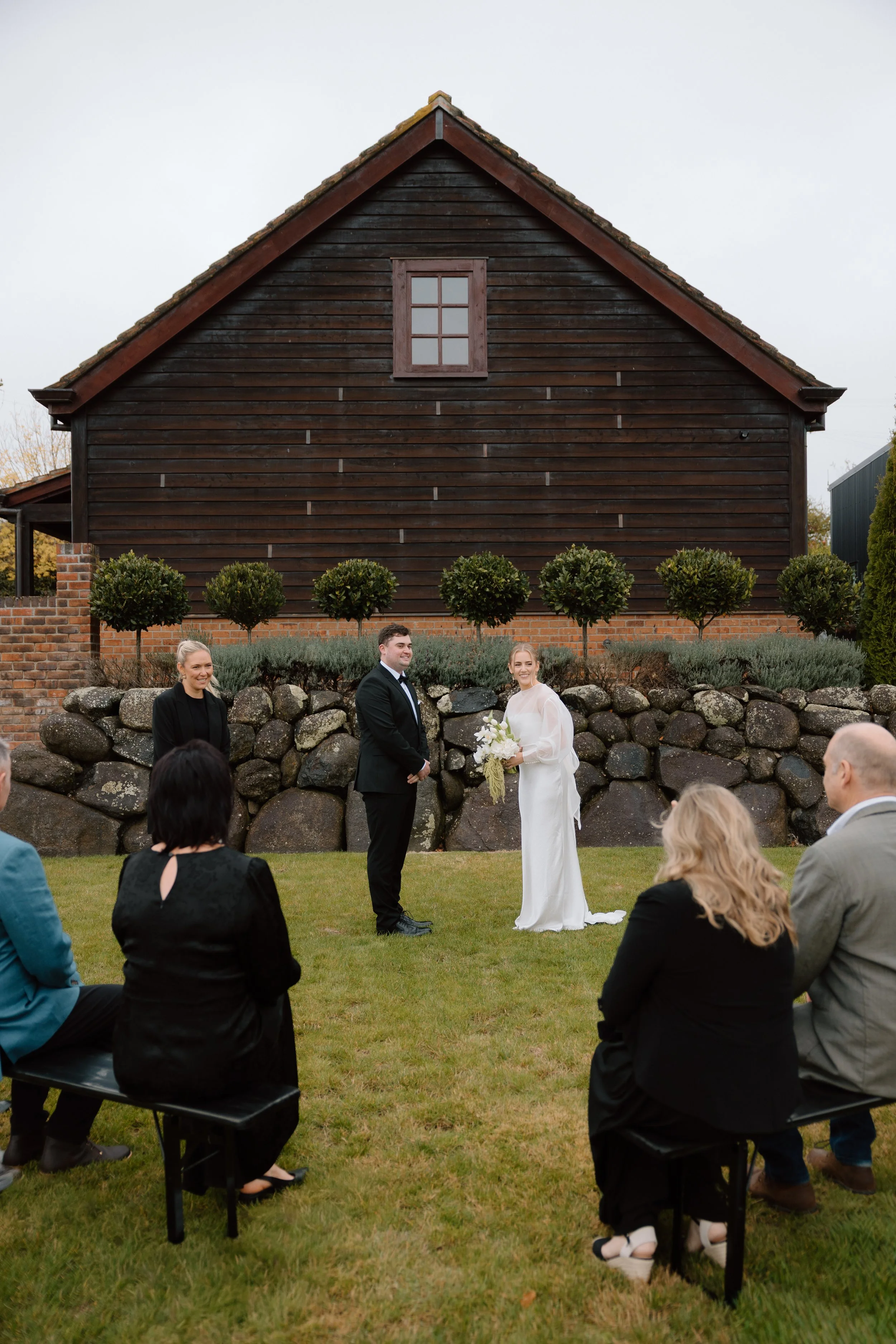 Outdoor wedding ceremony with a couple standing before an audience, in front of a large dark wooden building with a small window, surrounded by greenery and rocks.