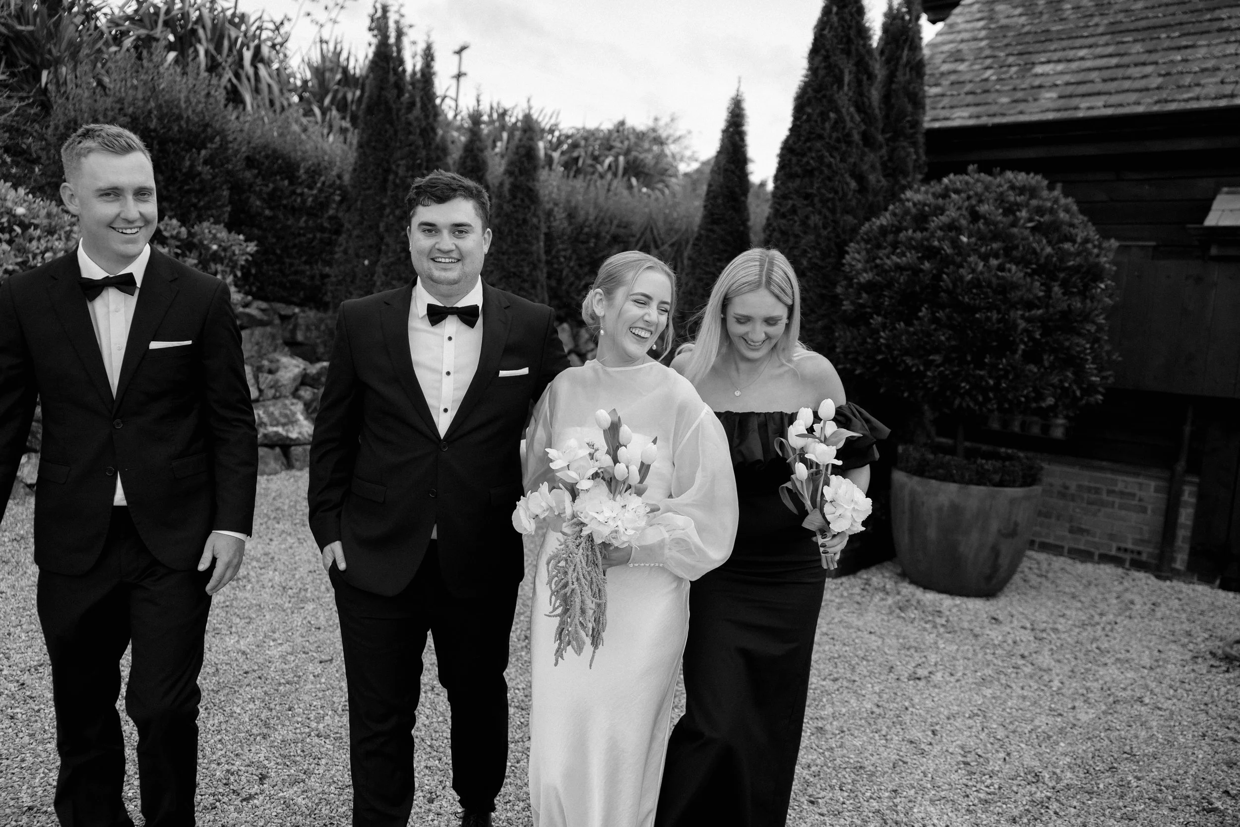Black and white photo of four people dressed in formal attire, standing outdoors with trees and bushes in the background. The woman in the center is holding a bouquet of flowers and wearing a wedding gown, smiling with her eyes closed.