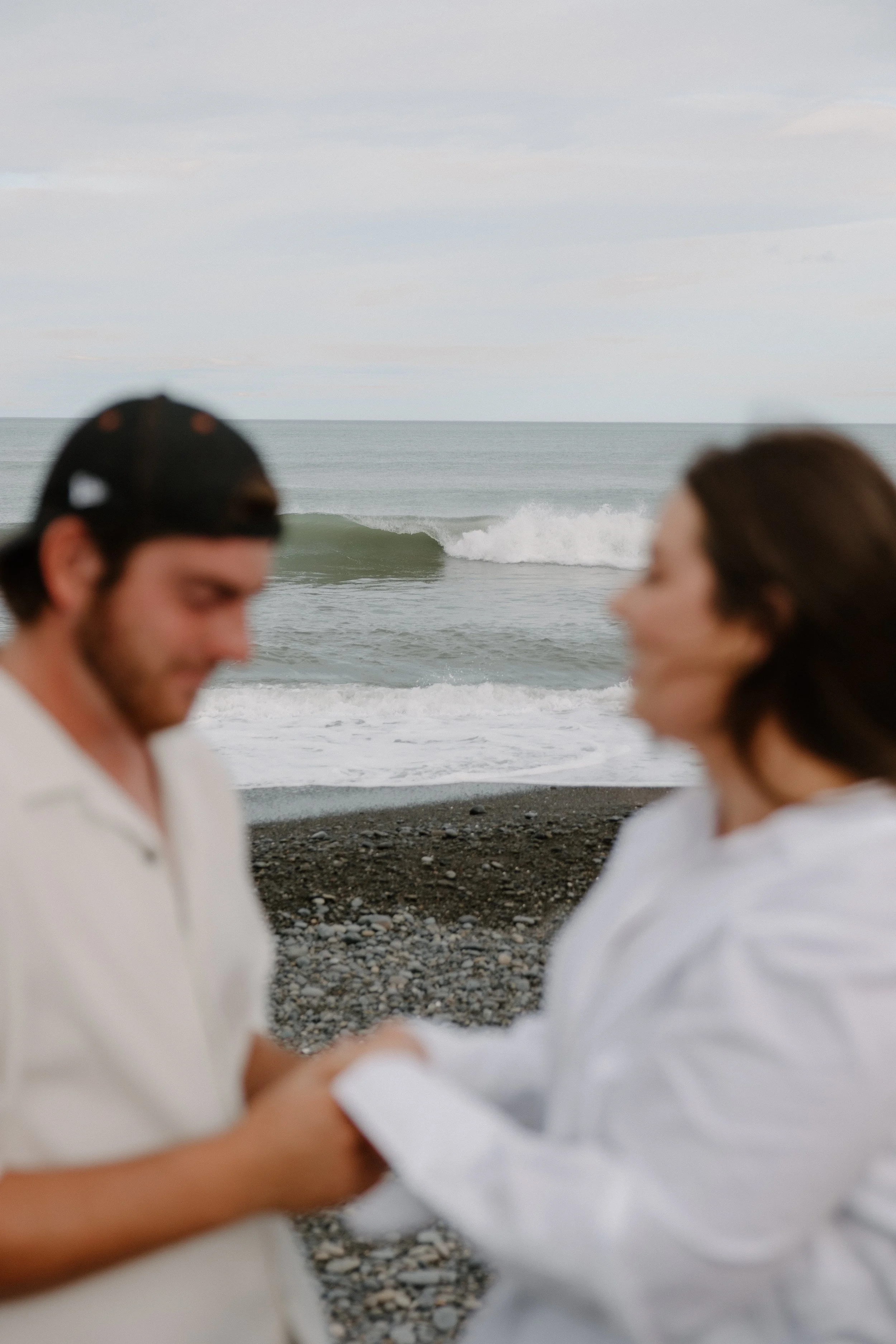 Two people standing on a rocky beach in white clothing, with an ocean and cloudy sky in the background.
