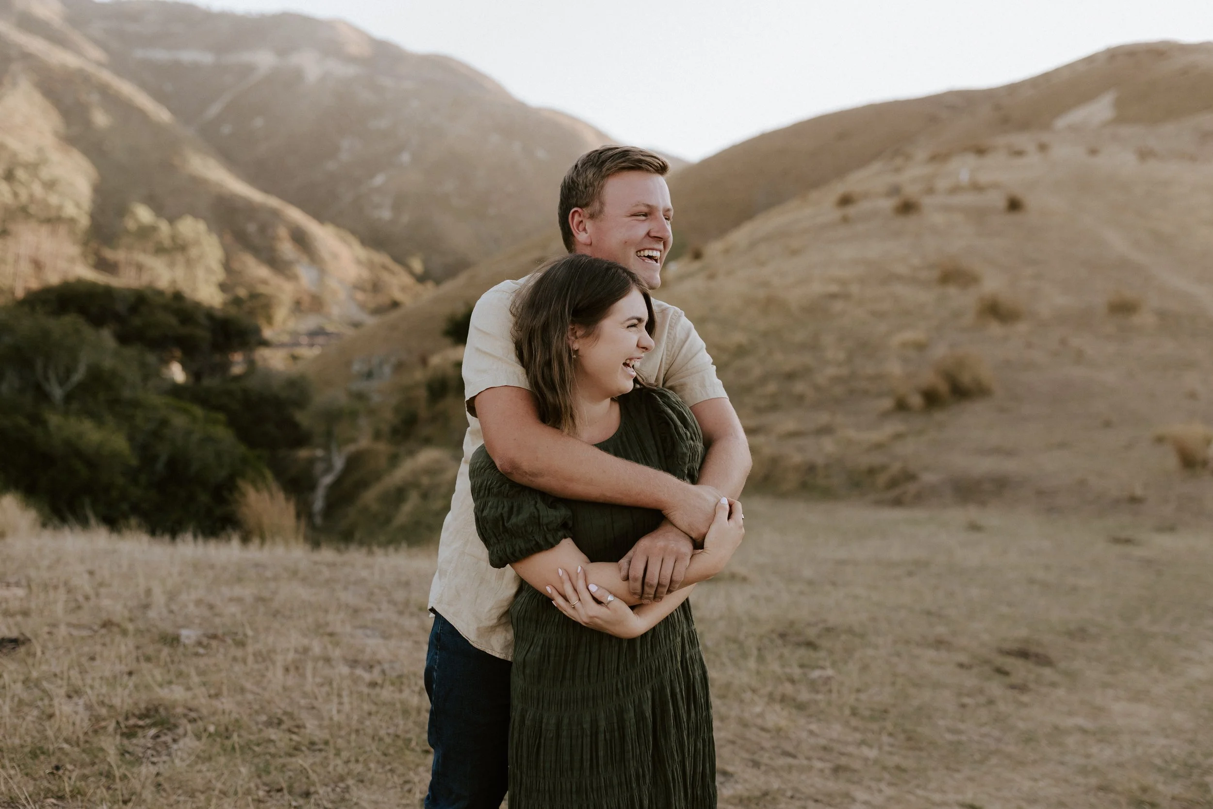 A couple standing outdoors, the man hugging the woman from behind, both smiling and laughing in a scenic landscape with hills and sparse trees.