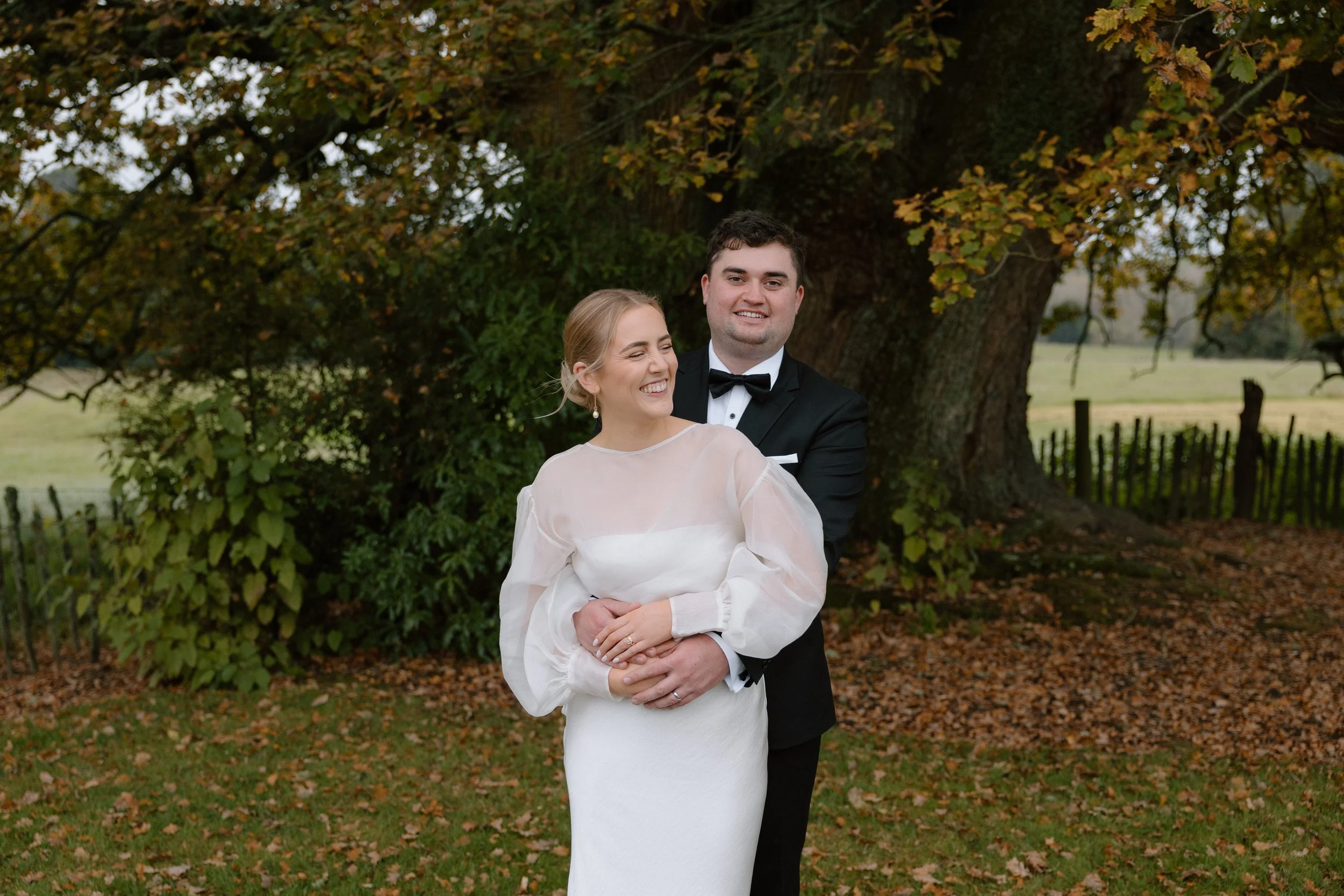 A newlywed couple standing outdoors in front of a large tree with autumn leaves. The bride is smiling with her eyes closed, wearing a white wedding dress with sheer long sleeves. The groom is behind her, smiling, wearing a black tuxedo with a bow tie