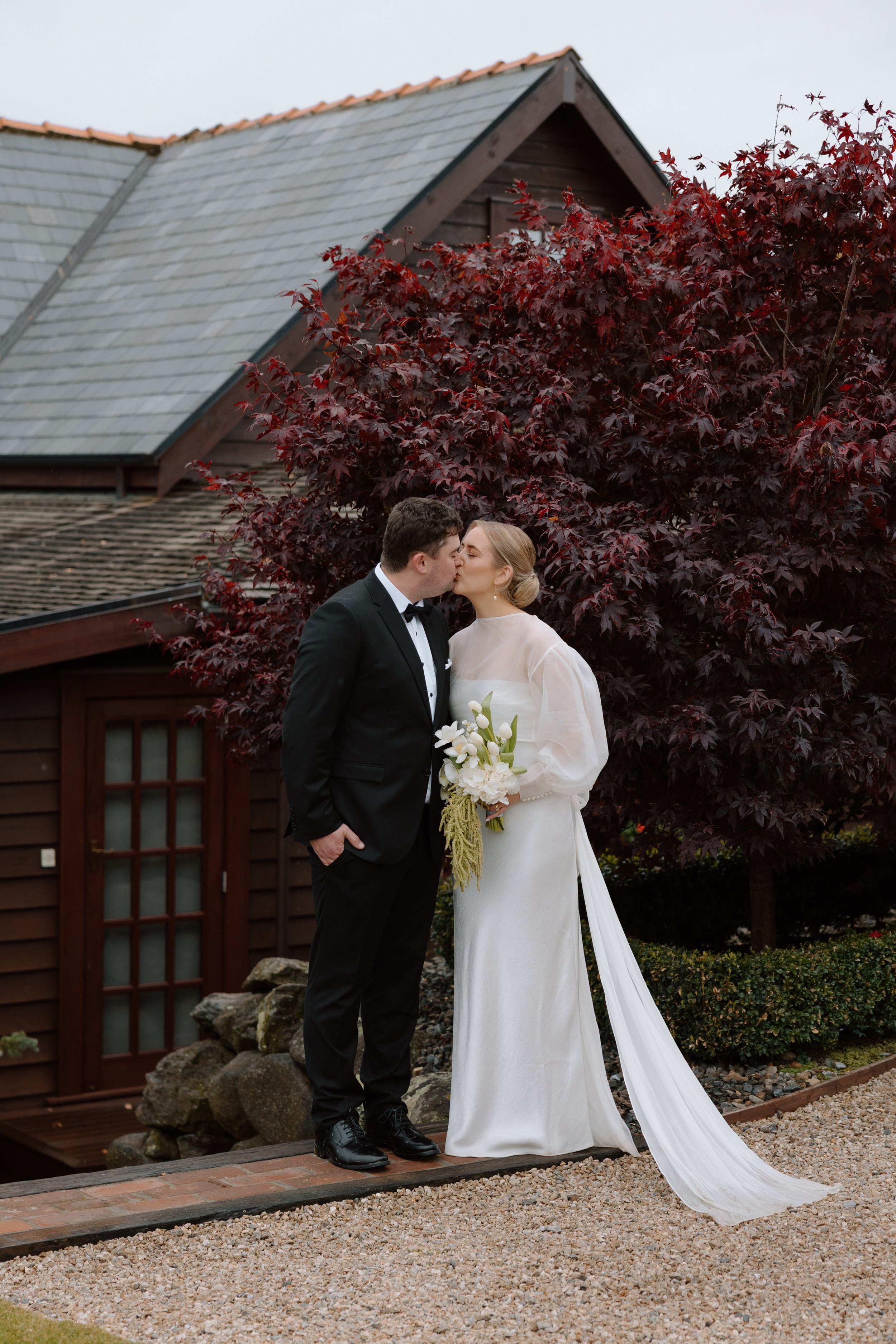 A newlywed couple sharing a kiss outdoors in front of a house with a red-leafed tree.