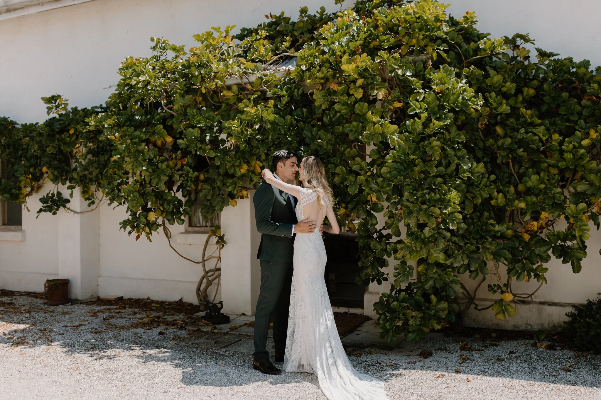 A couple in wedding attire sharing a romantic moment outdoors in front of a large green shrub, with the woman wearing a long white dress and the man in a dark suit.