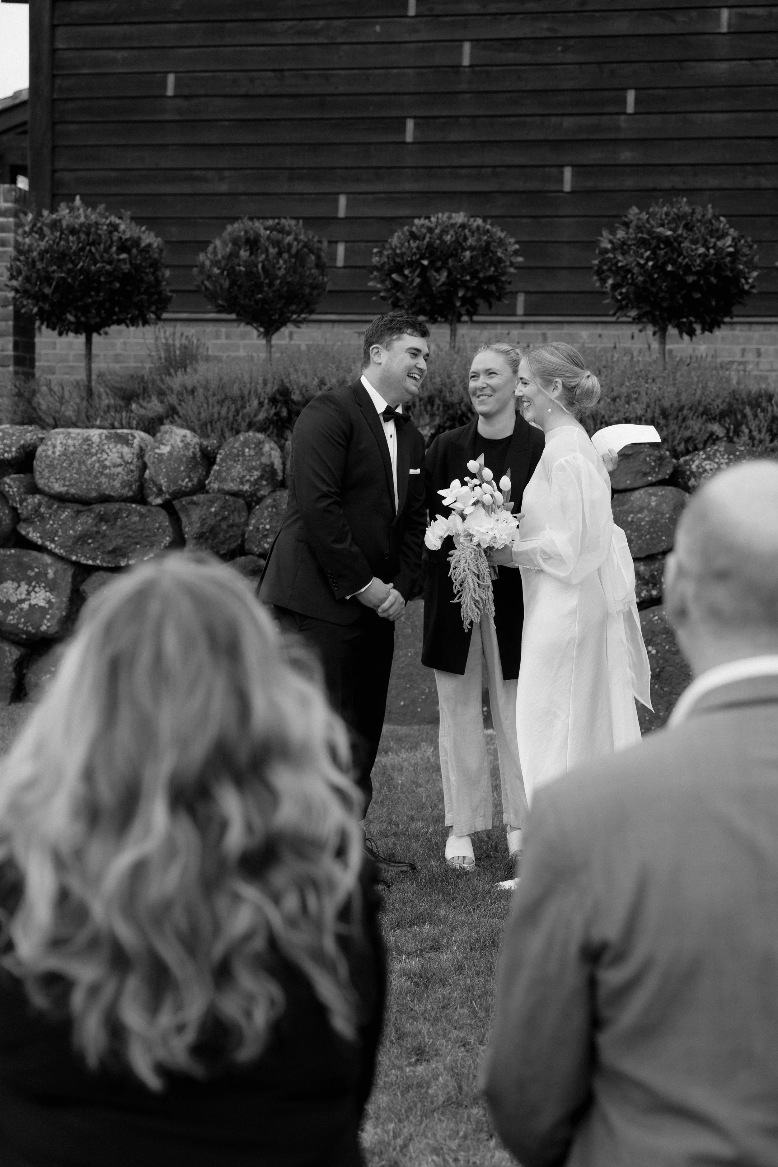 A couple getting married outdoors, standing face to face and smiling at each other, with an officiant holding a paper standing between them. The wedding is taking place in front of a stone wall with decorative trees, and guests are visible in the for