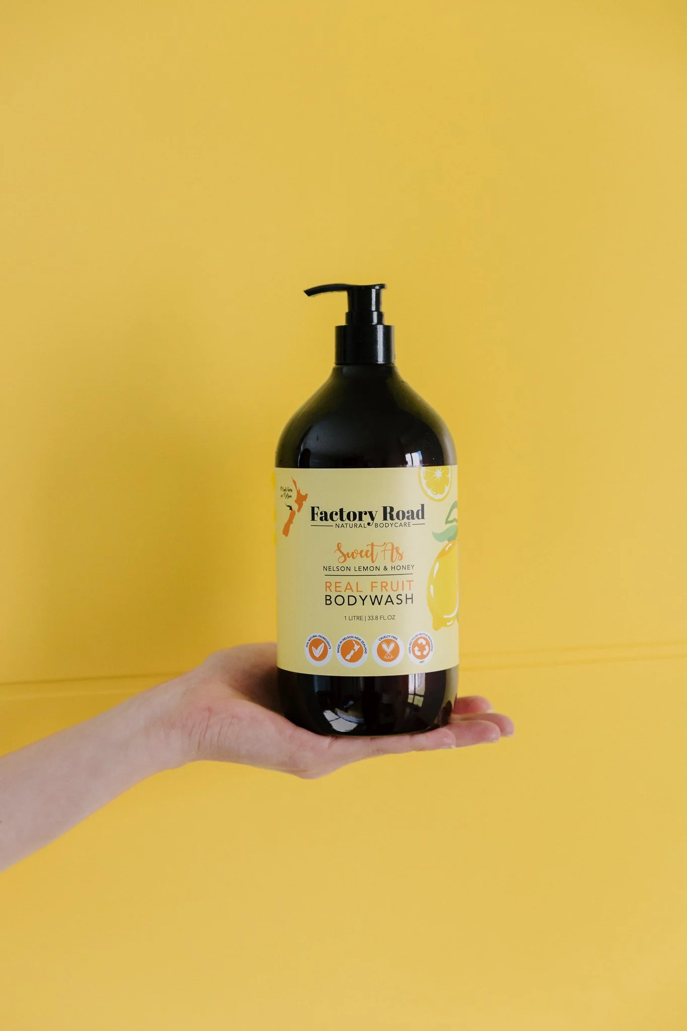 A hand holding a black bottle of Factory Road body wash with a yellow background. The label features lemon and honey imagery and the text "Sweet As Nelson Lemon & Honey".