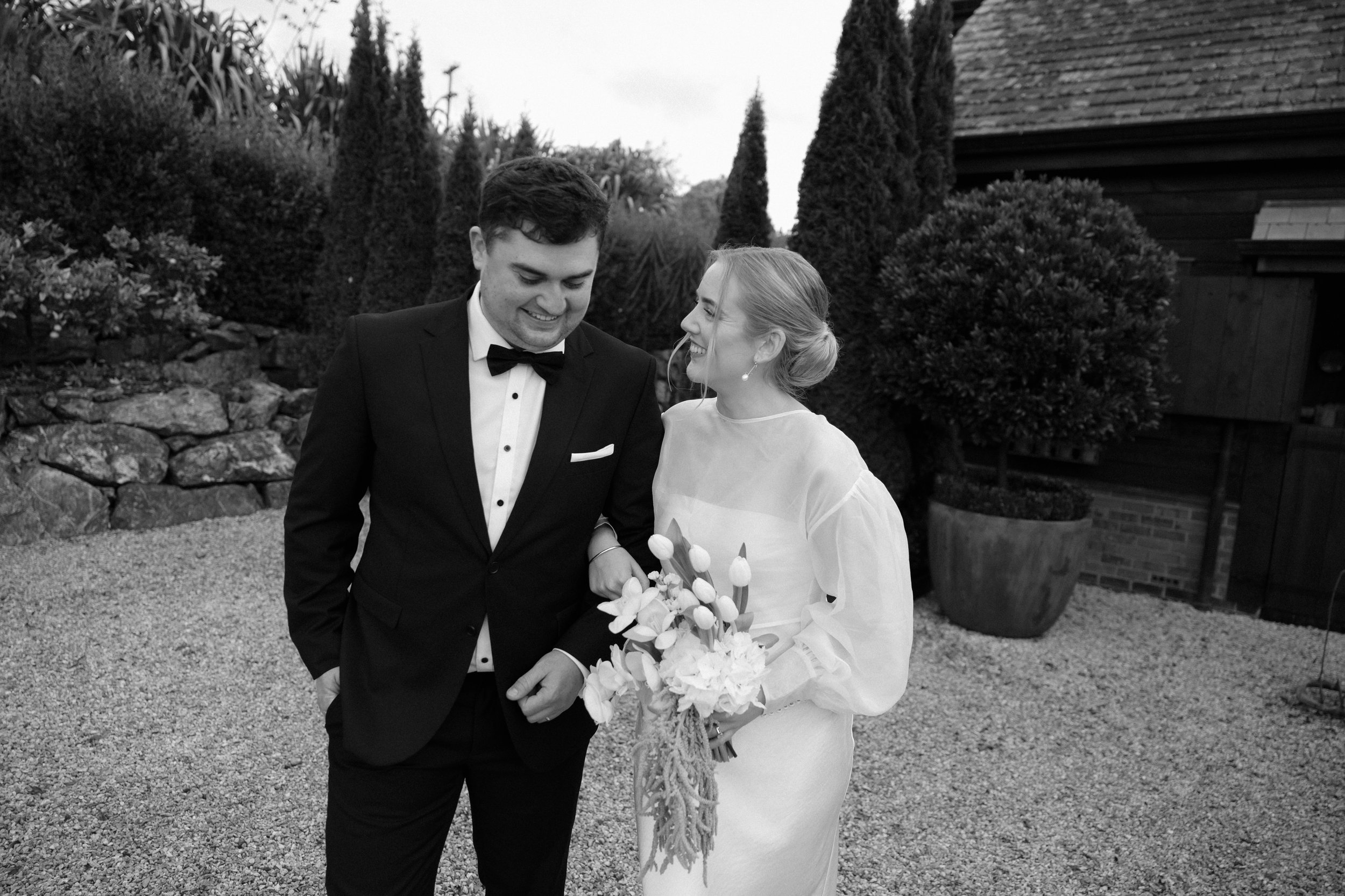 A black and white photo of a bride and groom smiling at each other, outdoors in a garden setting with trees, bushes, and a building in the background. The bride holds a bouquet of flowers.