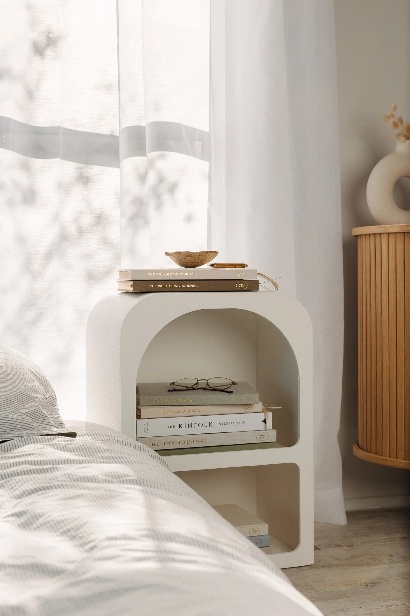 A cozy minimalist bedroom corner with a white arched shelf holding a stack of books, a pair of glasses on top, a small bowl, and a gold pen. A white zigzag-patterned bedspread is partially visible in the foreground. To the right, a round wooden side table with a vase of dried flowers and a white circular sculpture. Behind, sheer white curtains let in soft natural light.